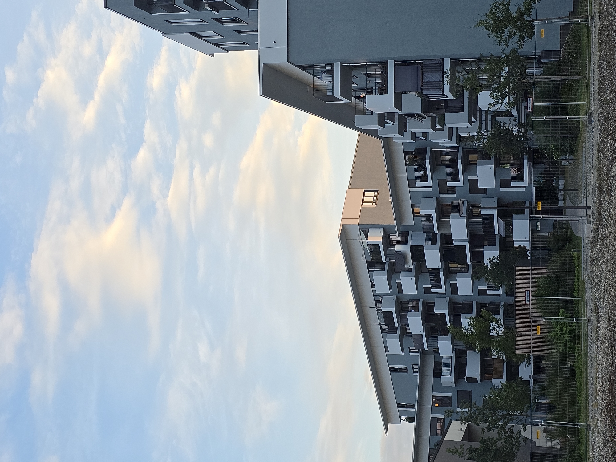 This is a photograph of a modern apartment complex in Munich, Germany, taken during the day. The sky is a light blue with wispy clouds. The buildings are grey with white accents and feature numerous balconies with black railings. Trees and a fence are visible in front of the buildings. The foreground shows a gravelly area. There are no people visible in the image. The architecture suggests a residential area, possibly a new development.