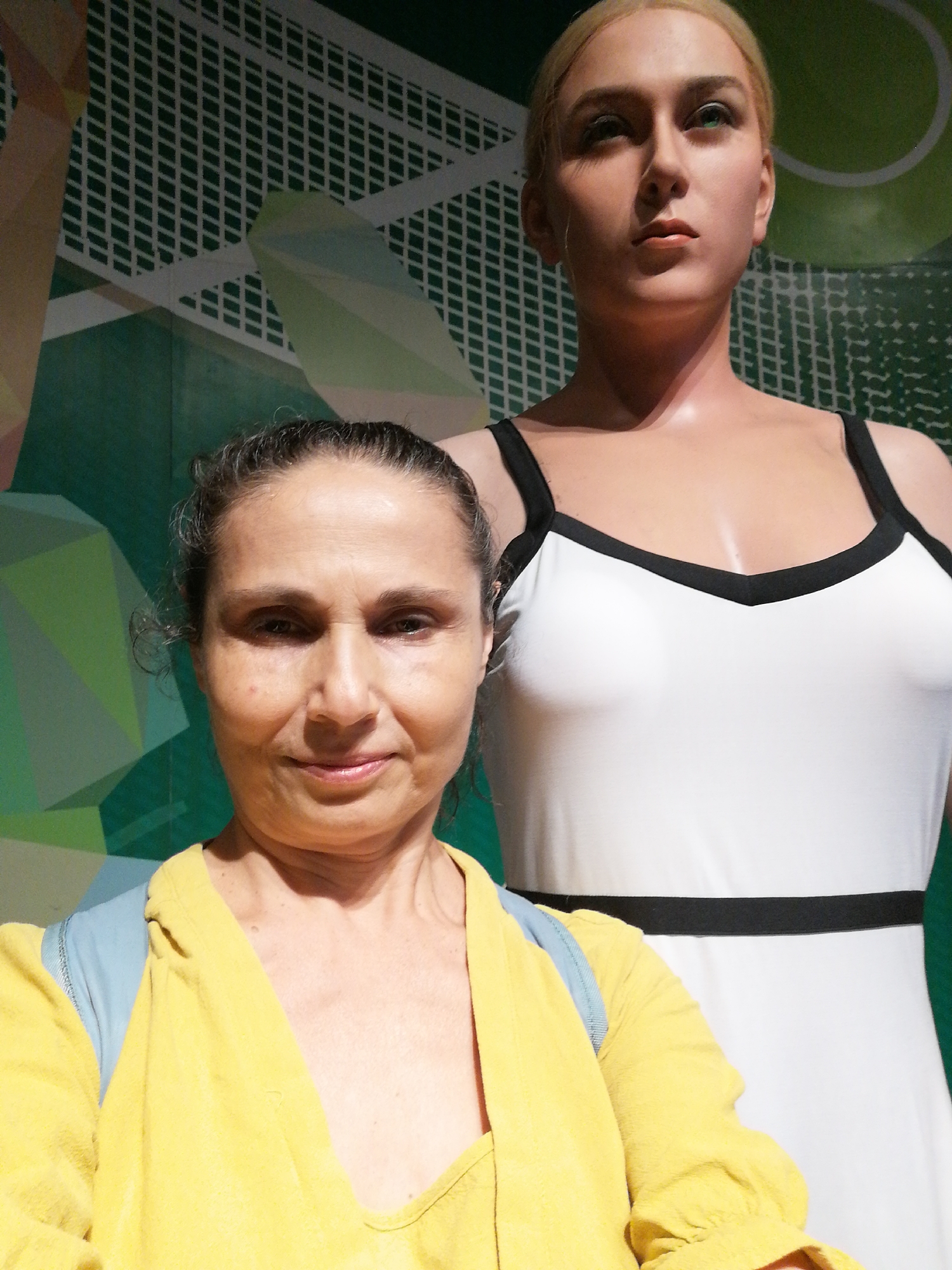 A medium shot depicts a woman in a yellow top next to a wax figure of a light-skinned woman in a white dress with black straps and a black band at the waist. The background shows a graphic design of green and white. The location is Unknown, Unknown.