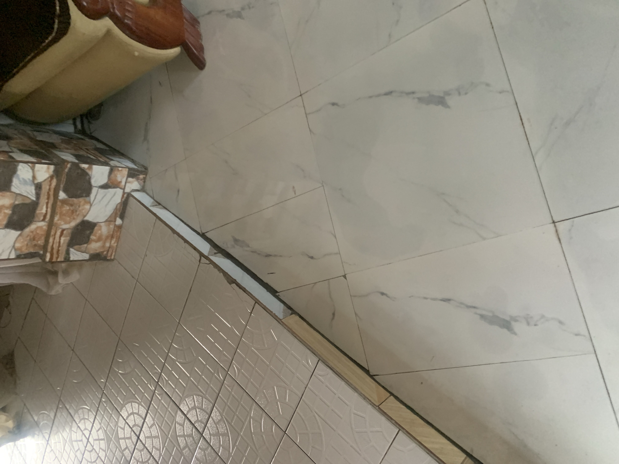 The image shows an interior floor with two different types of tiling. On the left, there is a section of white square tiles with a raised circular pattern. This tiled area appears to be raised slightly, with exposed wooden edging. To the right, the floor transitions to larger, rectangular white tiles with grey marble-like veining. In the background, a portion of a decorative tiled wall and the side of a light-colored sofa with a wooden leg are visible. The scene depicts an indoor setting, likely a home or common area. The location within the city of Jauro Yinu, Nigeria, is not discernible from the visual information provided in the image.