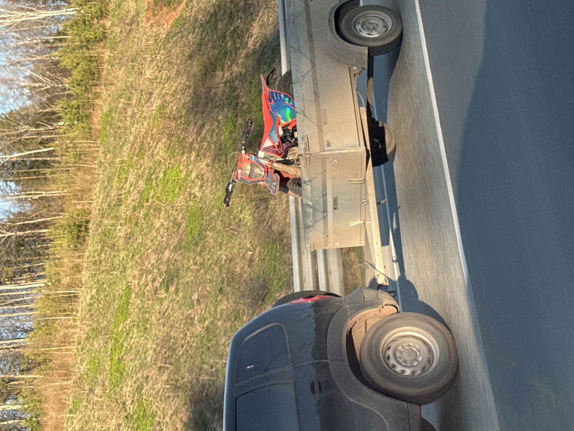The image shows a black van with an open trailer on a highway near a grassy area with trees, likely a highway shoulder. A red and blue ATV is loaded onto the trailer. The location is not definitively confirmed to be in Zvyozdny, Russia; this cannot be ascertained from the image alone. There are no people visible in the image.