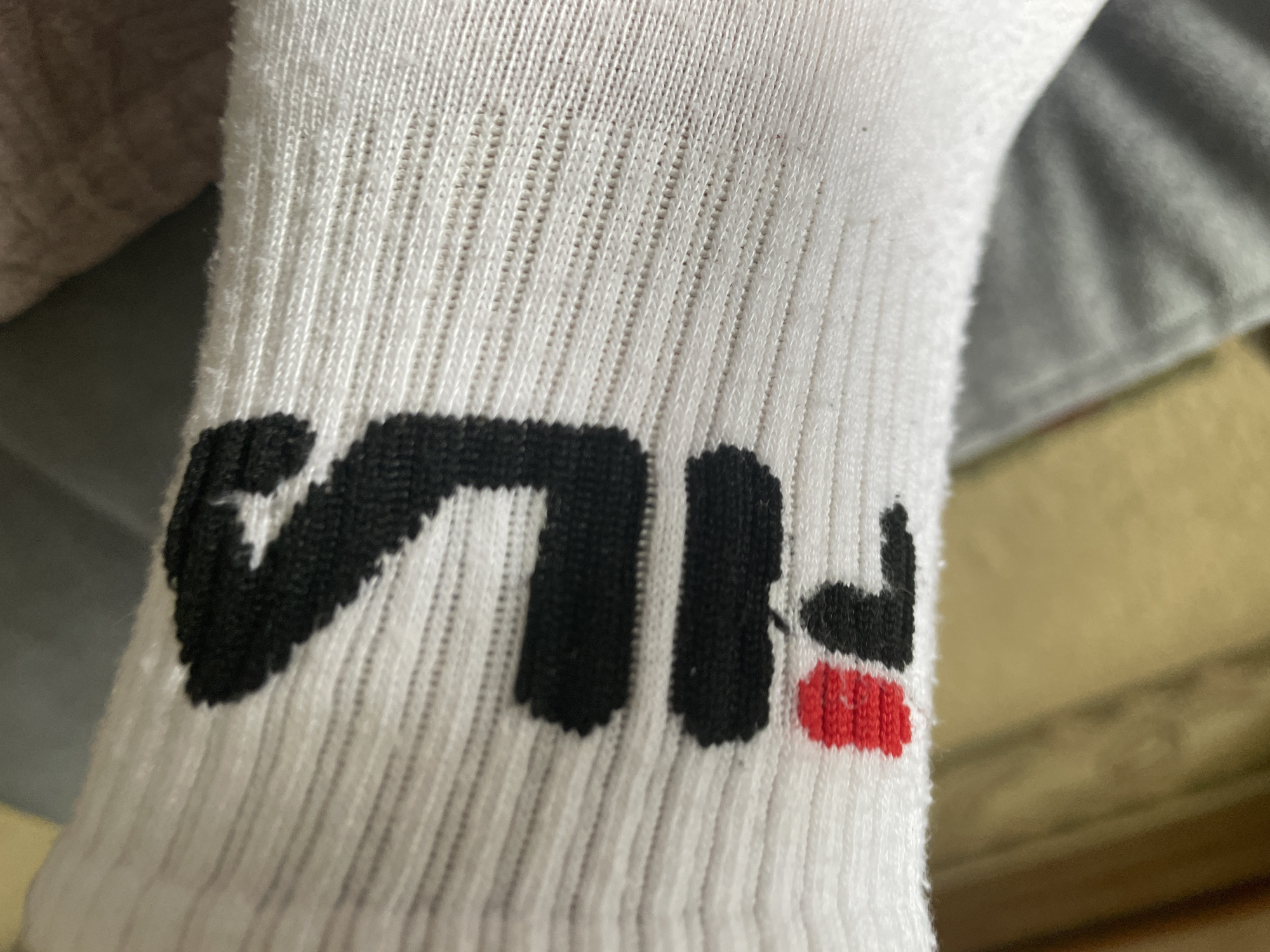 A close-up shot shows the Fila logo printed in black and red on a white ribbed sock. The sock is likely being worn, as it appears to be stretched over a foot or ankle. The background is out of focus, but appears to be a soft surface, possibly fabric or upholstery. The lighting is natural and appears to be coming from the side. The image is crisp and well-exposed, allowing for a clear view of the Fila logo and the texture of the sock.