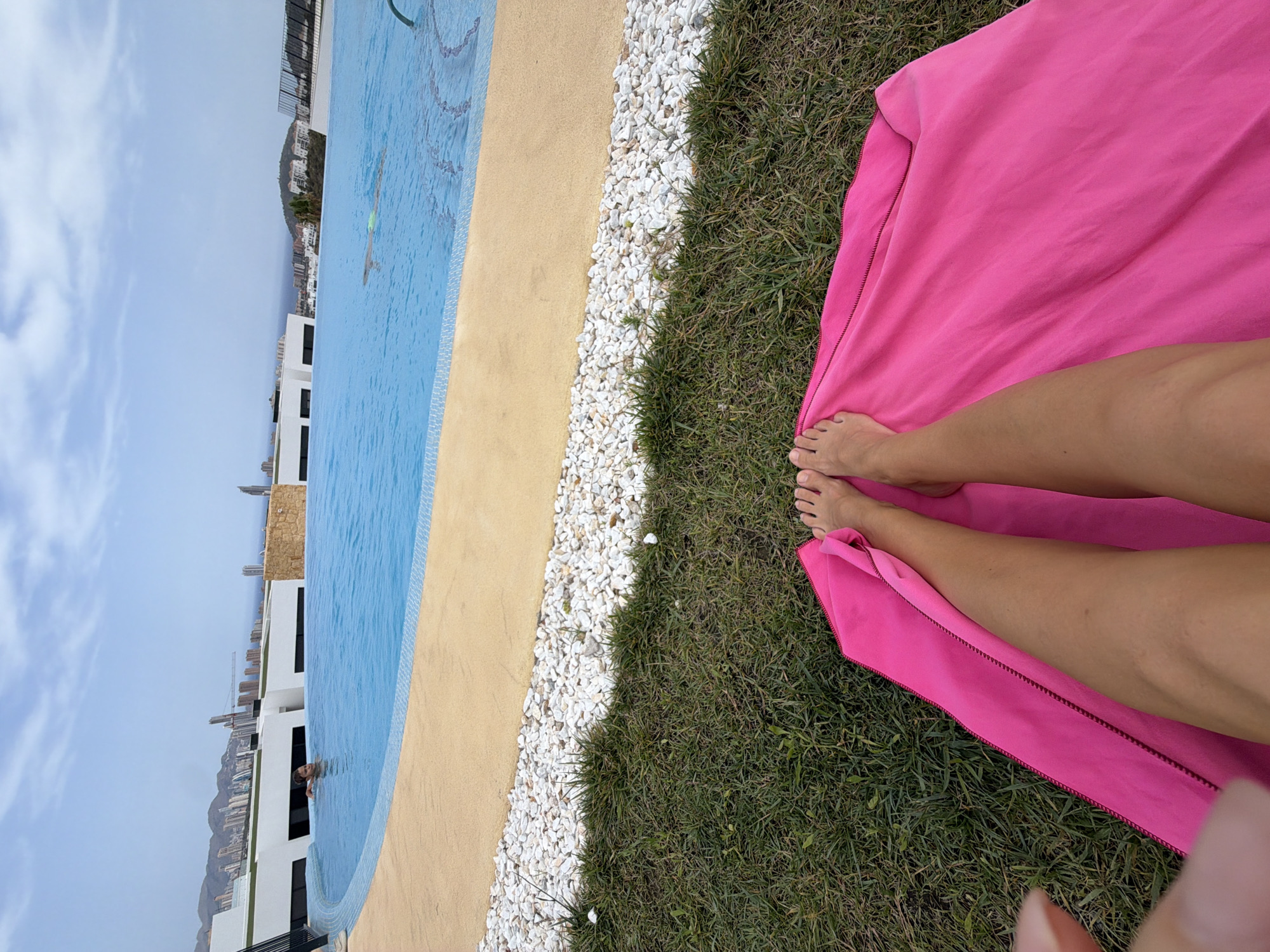 The image depicts a first-person perspective of a person resting on a bright pink towel spread on a grassy area. The person's legs, from the knees down, are visible, with their bare feet resting on the towel. In the background, there is an infinity swimming pool with bright blue water. Beyond the pool, a beige-colored pathway is visible, bordered by white pebbles. Further in the distance, a cityscape with tall buildings and mountains under a cloudy sky is present. The scene suggests an outdoor leisure setting, possibly a hotel rooftop or a private residence in Finestrat, Spain, given the architectural style and the coastal mountain backdrop. Two individuals are partially visible in the pool, one near the edge and another further out.