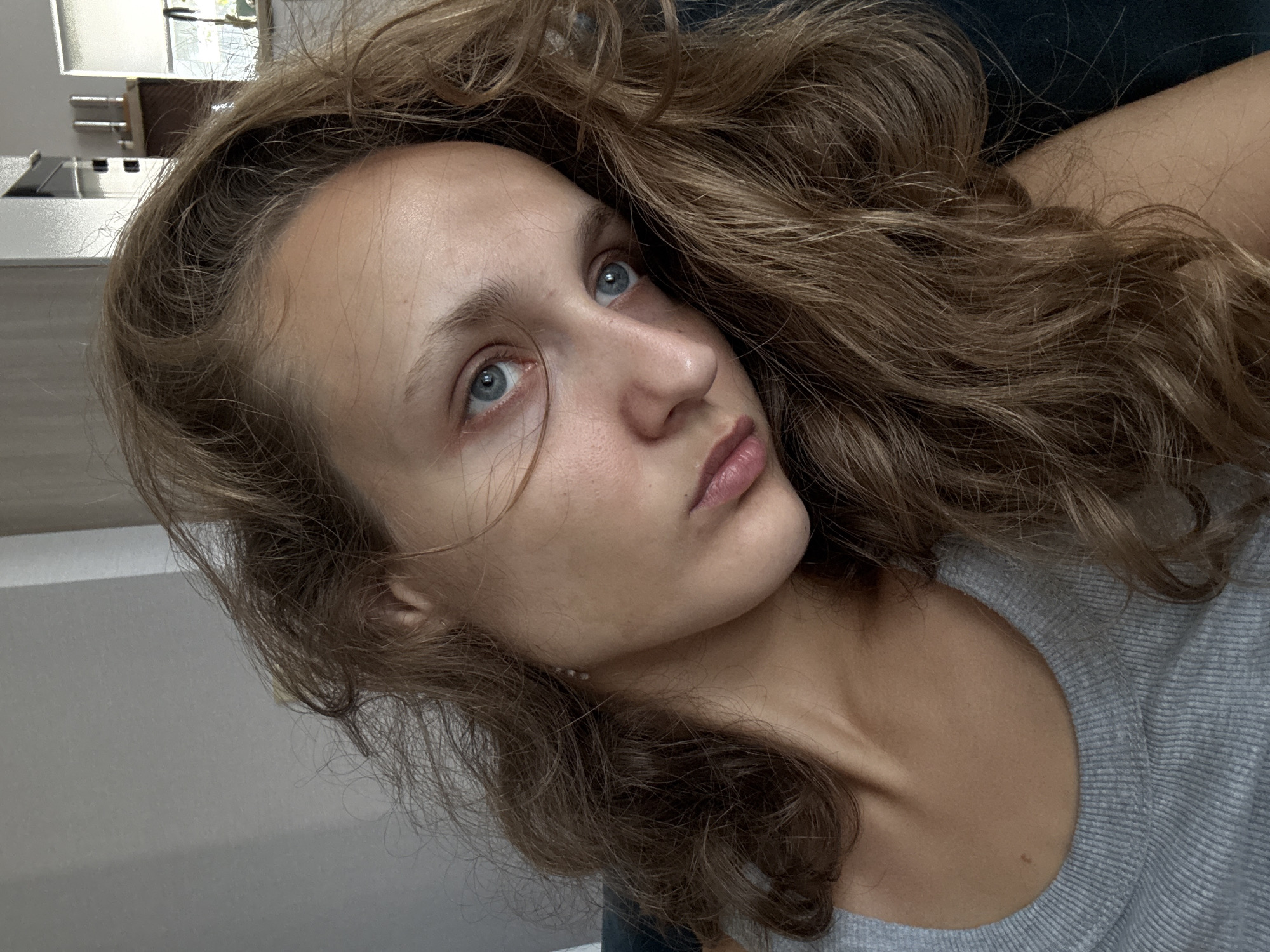 The image is a close-up, selfie-style photograph of a young woman with light brown, wavy hair and blue eyes. She is wearing a grey ribbed tank top. Her head is tilted, and she is looking off to the side with a somewhat pensive or melancholic expression. The background appears to be an indoor setting, possibly a living room, with a light-colored wall and a window with blinds visible in the upper right corner. The lighting is natural and appears to be coming from the side. There are no other people or animals visible in the image. The woman has a mole on her collarbone. No specific landmarks or elements indicative of the city of Подшивалово, Russia are present in the image.