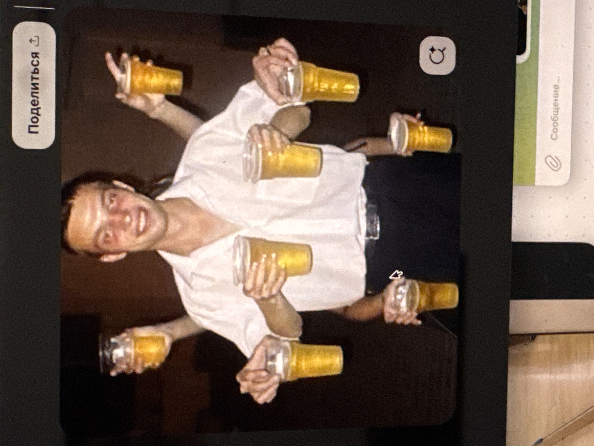 This is a medium shot, low-angle, and slightly blurry photograph of a person being held up by multiple arms, each holding a clear plastic cup filled with a yellow, bubbly liquid, likely beer. The person appears to be male, with a wide, excited smile and flushed cheeks, suggesting a celebratory or playful atmosphere. He is wearing a white t-shirt. The background is dark, making the lit subjects stand out. The overall impression is one of revelry and camaraderie. There is some text visible on the left side of the image which says "Поделиться" which is Russian for "Share". There is also a small icon of an arrow pointing upwards, also a share icon. On the right side of the image, there is a light green strip with text that says "Сообщение" which is Russian for "Message". There is also a paperclip icon, suggesting this might be an image shared within a messaging application. Given the context of Kuta Selatan, Indonesia, this could be a scene from a beach party or a lively evening gathering in a popular tourist area known for its nightlife. The lighting and overall mood suggest it is likely nighttime or indoors with dim lighting.