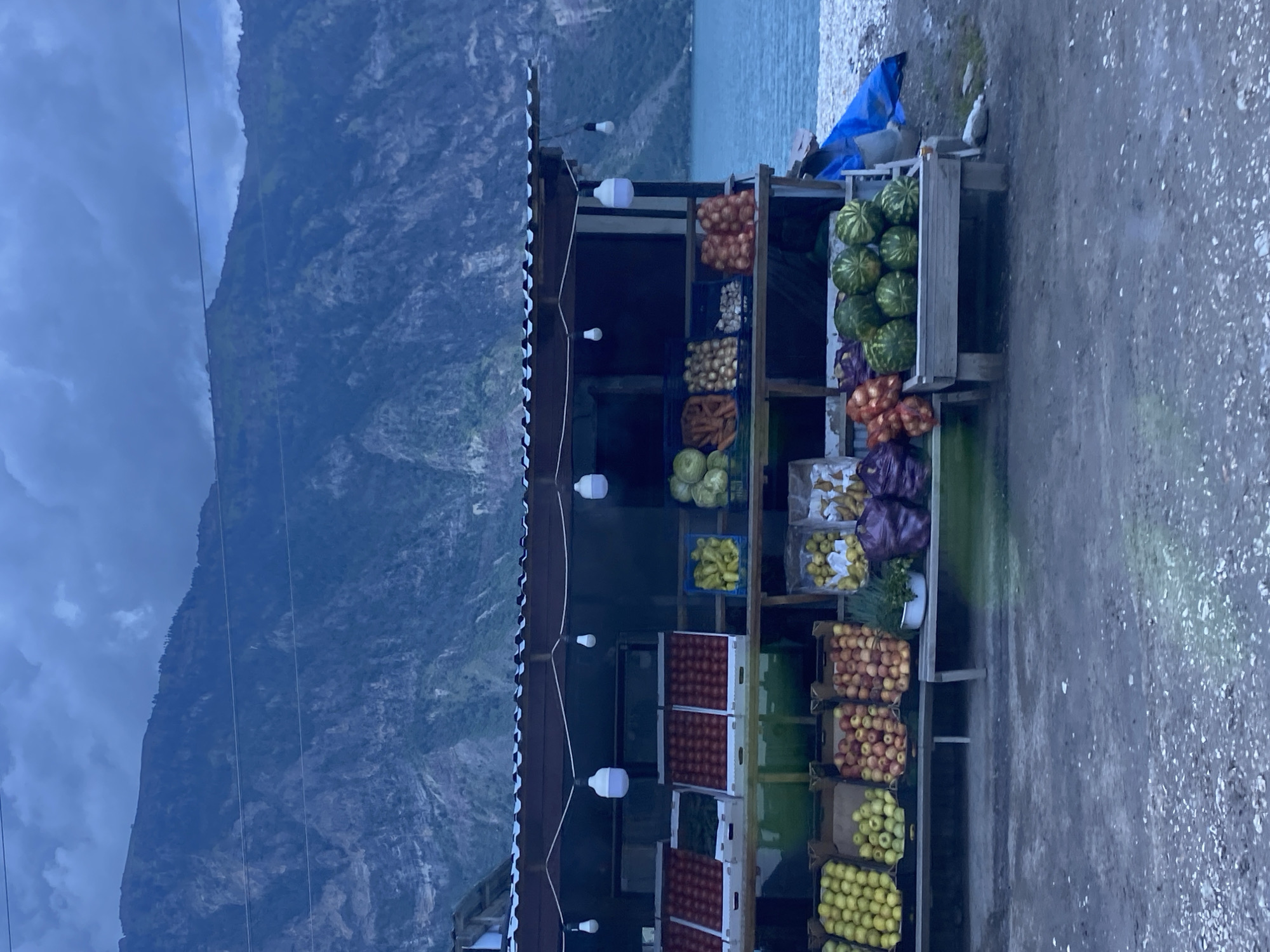 A roadside fruit and vegetable stand is set up in front of a body of water with a mountainous backdrop. The stand has a corrugated metal roof supported by wooden posts, and string lights with bare bulbs are hung across the front. Various fruits and vegetables are displayed in wooden crates and burlap sacks, including apples, watermelons, onions, and carrots. The ground in front of the stand is covered with gravel and dirt. In the background, a large, calm body of water stretches to the horizon, and behind it, imposing mountains rise, their slopes covered in vegetation and rock faces. The sky is overcast, with dark, heavy clouds suggesting the possibility of rain or an approaching storm. There are no people visible in the image, and the scene appears to be unattended. The overall impression is one of a remote, rustic marketplace situated in a dramatic natural landscape. The location is identified as Irganay, Russia.