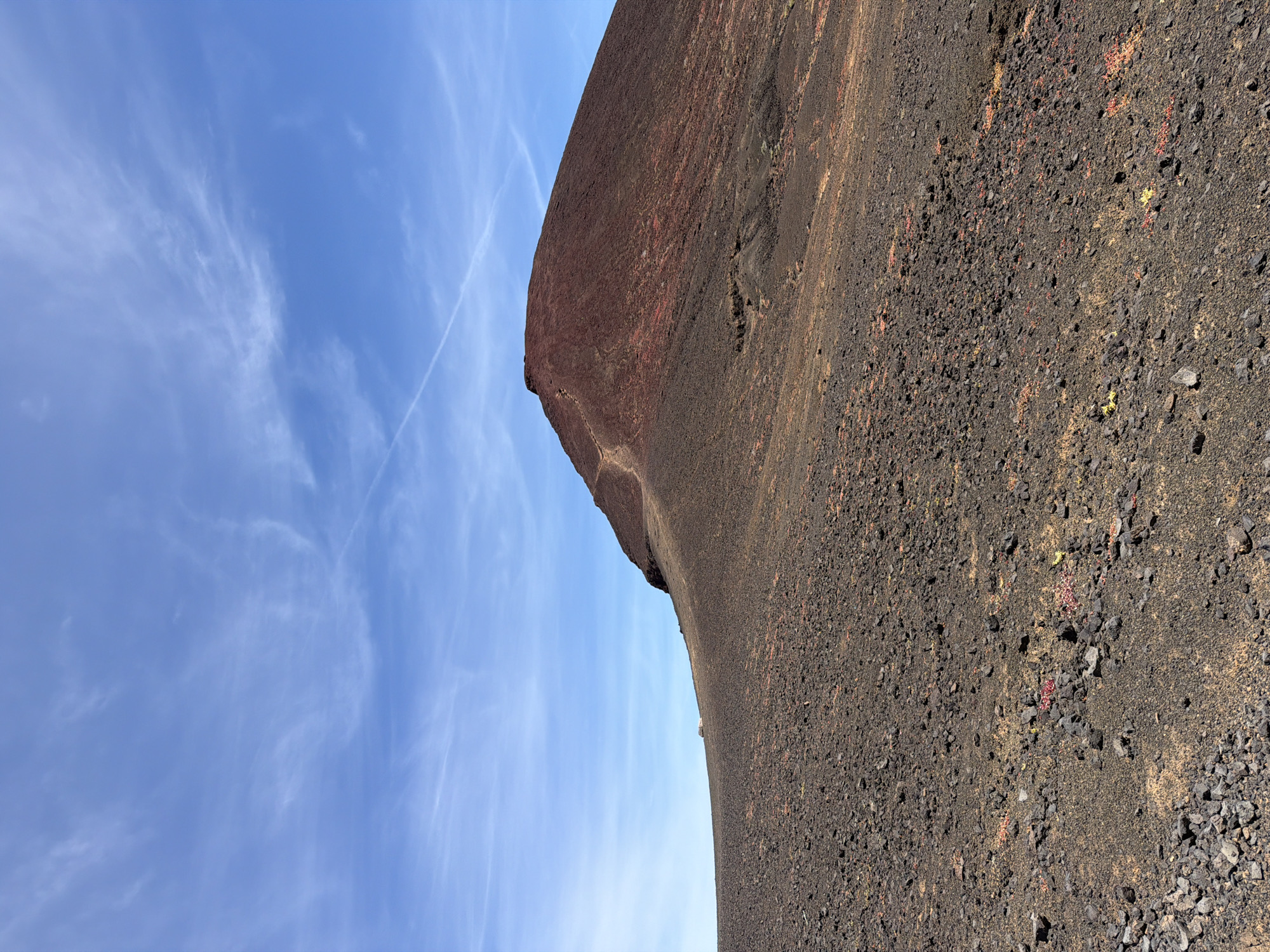 The image shows a landscape in Tinajo, Spain. On the right, a steep, dark brown and black volcanic rock formation rises from the ground. The surface is covered with small rocks and sparse vegetation. To the left, a blue sky with wispy white clouds provides a contrasting backdrop. The photograph appears to be taken from a low angle, emphasizing the height and ruggedness of the volcanic rock.