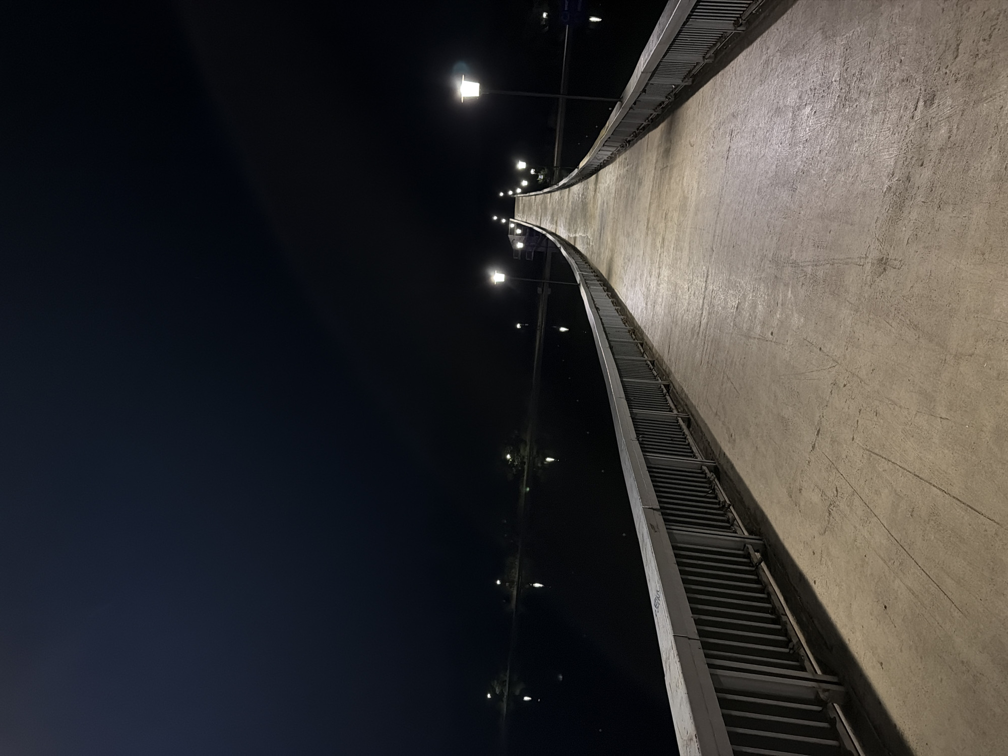 The image shows a concrete pedestrian bridge over water at night. The bridge is lined with metal railings and illuminated by lampposts. The water reflects the lights, creating a serene atmosphere. The sky is dark and clear. The location is identified as Plovdiv, Bulgaria. There are no people visible in the image.