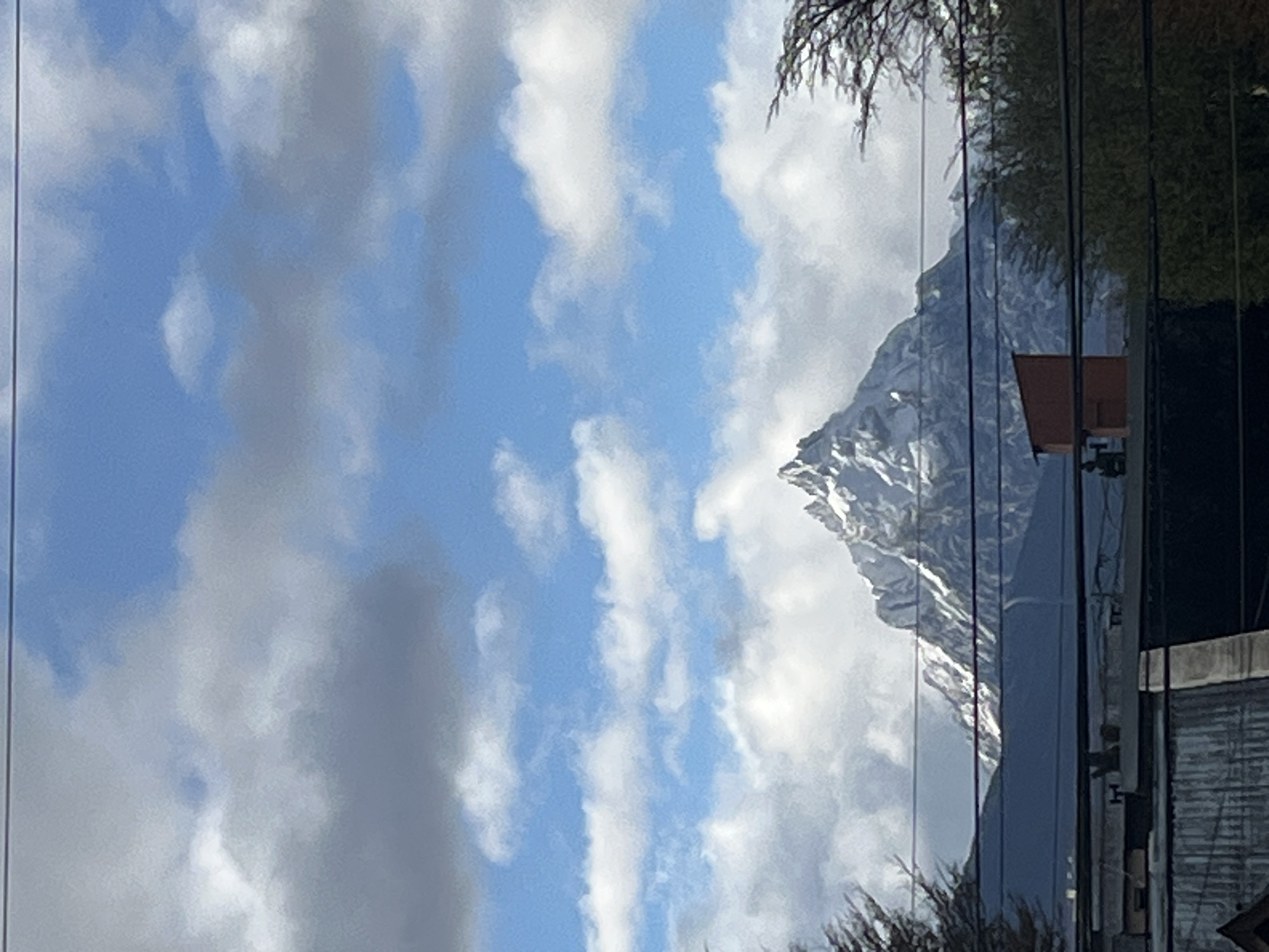 This image, taken in Ushuaia, Argentina, captures a striking view of a snow-capped mountain under a partly cloudy sky, seen through a reflective surface, likely a window. The sky dominates the upper and left portions of the frame (when correctly oriented), showcasing a clear blue punctuated by dynamic white and grey clouds.
A majestic, rugged mountain, covered in snow and rock, rises prominently on the right side of the image. Its sharp, jagged peaks indicate a formidable natural landscape. Through the glass, various reflections are visible, including parts of a reddish-brown structure, possibly a building or cabin, nestled against the mountain, and a darker, perhaps corrugated, surface towards the bottom right. Several dark, vertical lines, appearing to be cables or structural elements, traverse the frame, some reflecting the sky and adding to the layered effect. Sparse, bare tree branches are also discernible in the upper and lower parts of the image, either reflected or viewed through the glass.
The scene depicts a clear, bright daytime, suggesting good visibility of the surrounding landscape. The combination of the imposing mountain, the cloudy sky, and the reflective foreground elements creates a complex and layered composition. No activity or individuals are depicted, and there is no visible text.