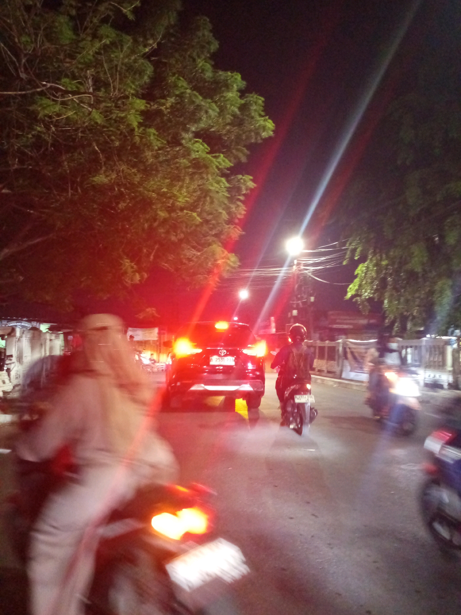 A blurry, night-time street scene in Bekasi, Indonesia shows several motorcycles and a car on a road. The image is dominated by red and white light flares from vehicle taillights and streetlights. In the foreground, a person on a motorcycle is blurred, wearing light-colored clothing. To the right of this figure, another motorcycle rider with a helmet is visible, also with a blurred appearance. In the middle ground, a black car with its brake lights illuminated is present, its license plate visible. Further down the road, more motorcycles are seen with illuminated headlights and taillights. Trees line the sides of the street, and streetlights are visible overhead. The overall impression is of a busy, active street at night, with motion blur contributing to the dynamic feel of the scene.