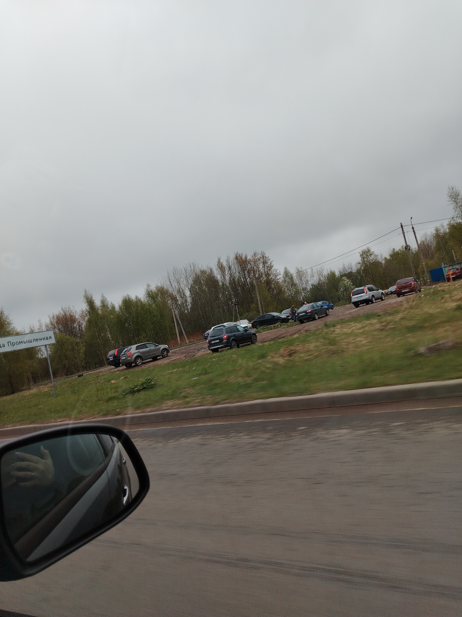 The image depicts a roadside scene in Заволжье, Russia, as viewed from inside a moving vehicle. The side mirror of the vehicle is visible in the foreground, reflecting the interior and the driver's hand. The road is asphalt, and the weather is overcast.
On the side of the road, there is a grassy embankment leading to a dirt lot where several cars are parked. Trees and power lines are visible in the background. A sign, partially obscured, reads "Промьшленная".