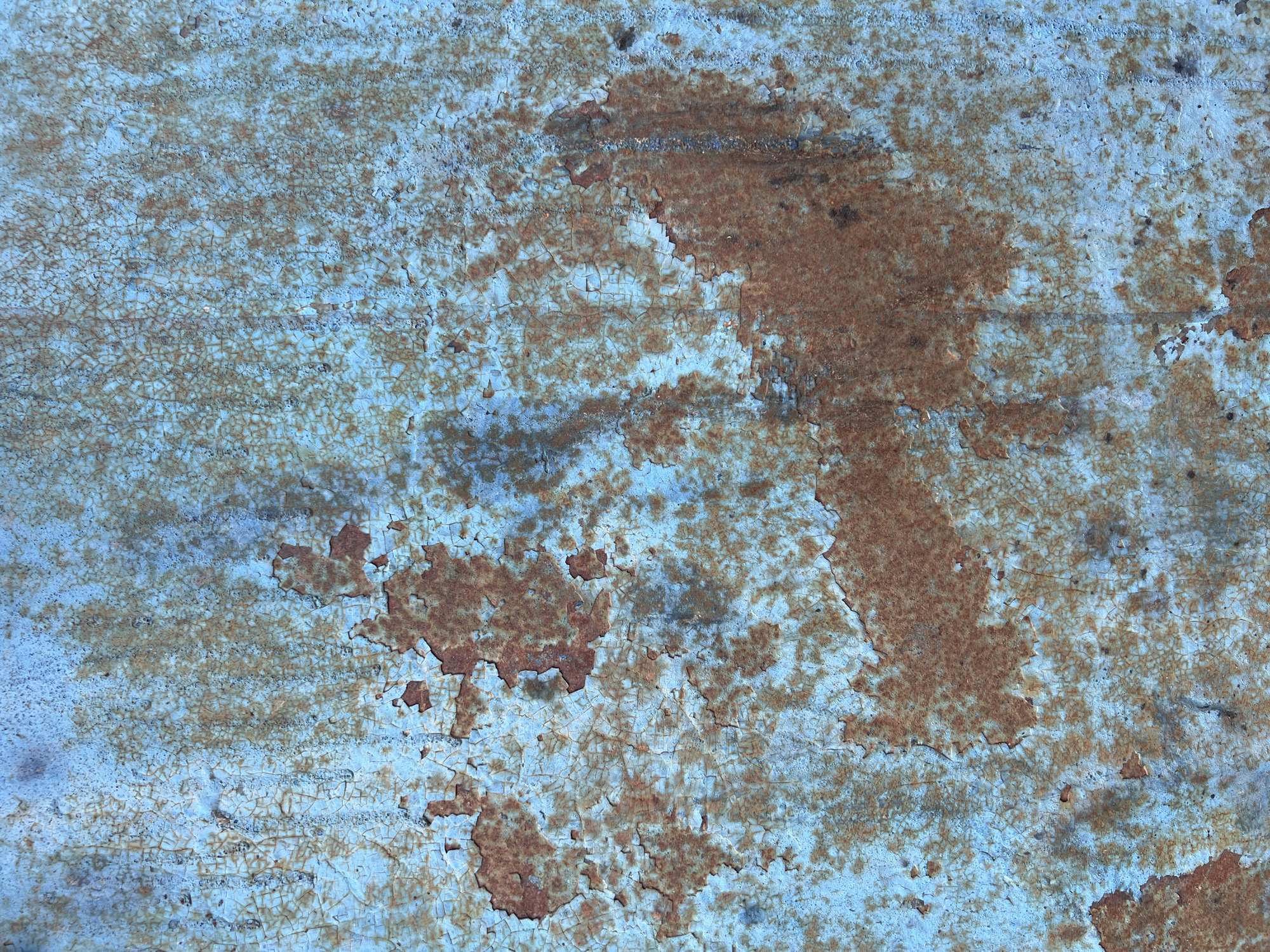 The image shows a close-up of a textured, weathered surface, likely metal, with peeling blue paint and visible rust. The paint is cracked and flaking in many areas, revealing a reddish-brown rust underneath. The texture is uneven and appears to be a close-up detail of an aged object. The image does not contain any information that would allow for the identification of a specific location within Moscow, Russia.