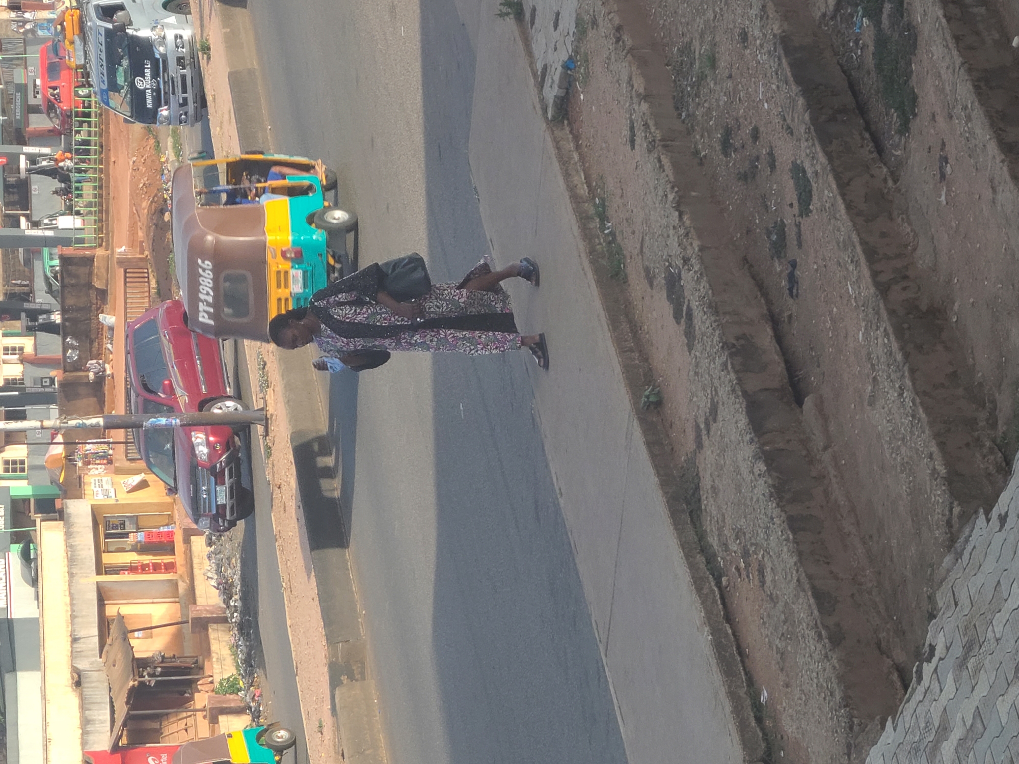 A woman is walking on a sidewalk in Gura Top, Nigeria, near a road with several vehicles including a “KWAYA ROSAR & SONS” minibus, a tricycle with the number “PT-19866”, and a red pickup truck. The area appears to be urban, with buildings and shops visible in the background. The sidewalk is adjacent to a bare patch of earth. The woman is wearing a patterned outfit and carrying a bag.
