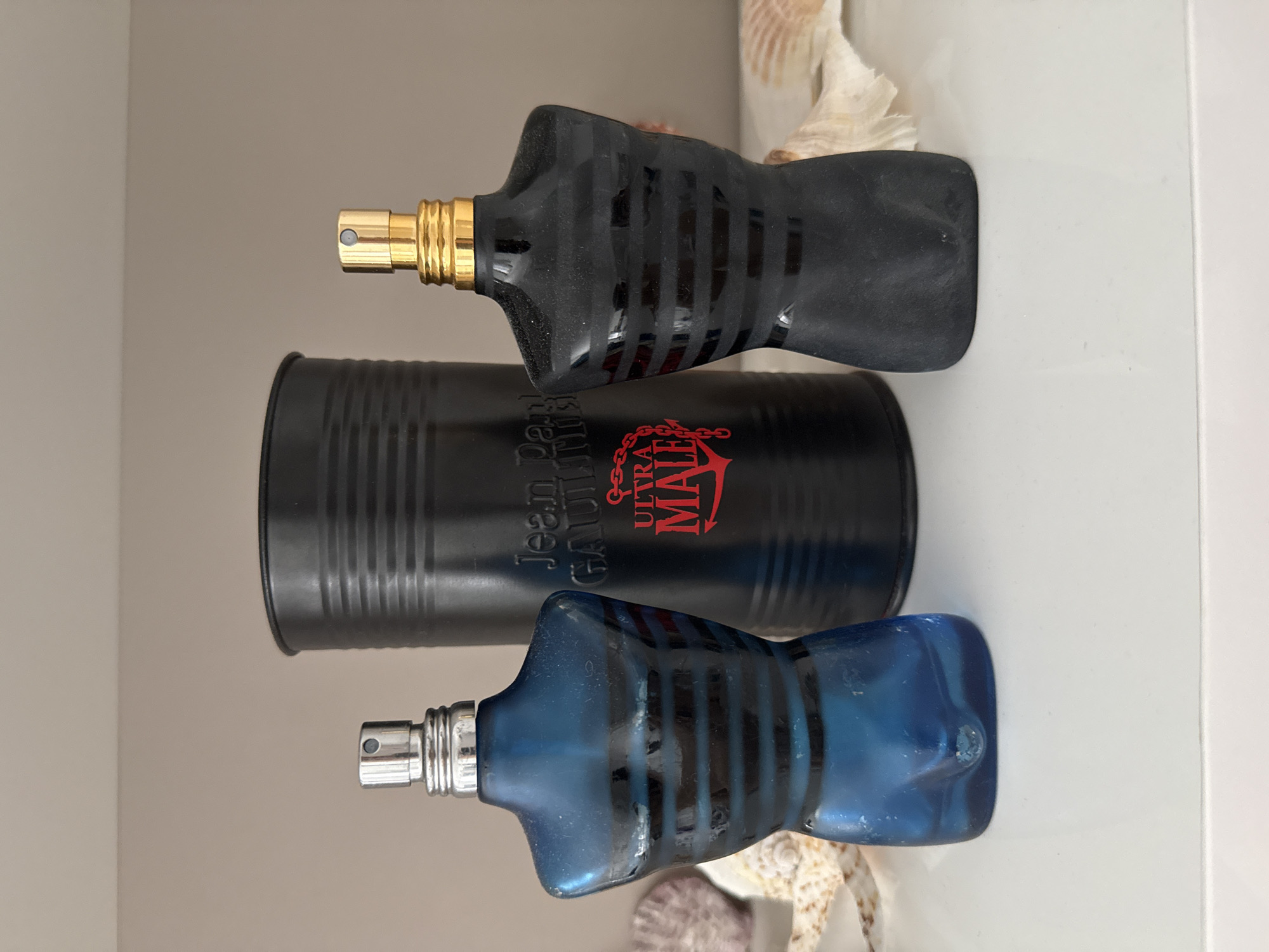 Two torso-shaped perfume bottles, one blue and one black, stand on a white surface. The blue bottle is on the left, and the black bottle is on the right. Behind them, a black cylindrical tin is visible, with the text "Jean Paul Gaultier ULTRA MALE" printed in red. The background is a plain beige wall. Seashells are placed on either side of the bottles. There are no people or animals in the image. The scene is indoors, likely a display on a shelf or table. The image does not provide any information about the city of Burgas, Bulgaria.
