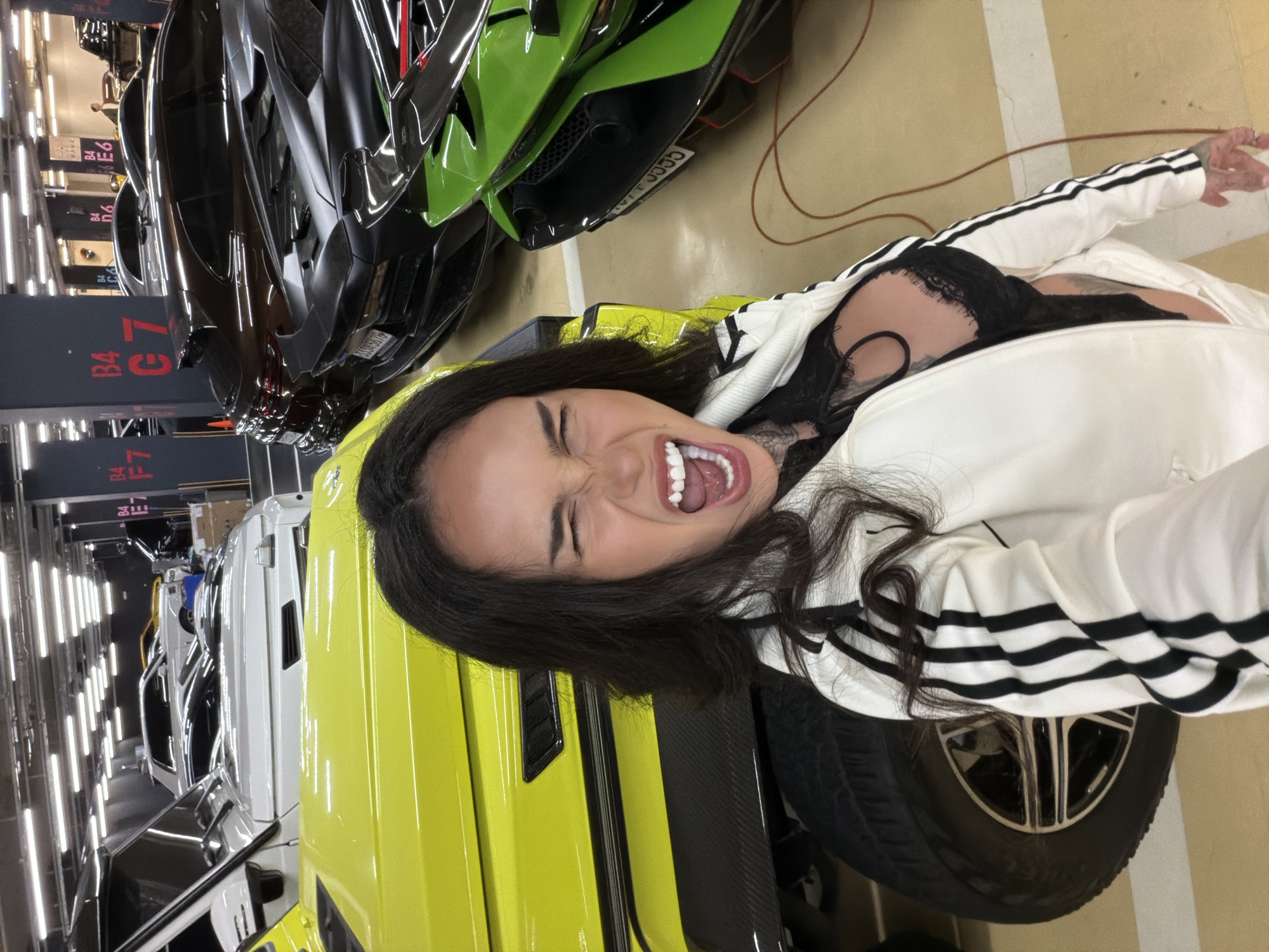 A rotated, high-angle close-up depicts a woman with long, dark hair wearing a white Adidas track jacket and black lace lingerie. She is on her back, mouth open, eyes squinted, and teeth visible. She is next to several luxury cars including a bright yellow Lamborghini and another dark car. The background includes a parking garage setting with overhead fluorescent lighting and signage indicating parking levels. Based on the visual cues, the location is likely in a high-end district of Seoul, South Korea, where luxury vehicles are common.