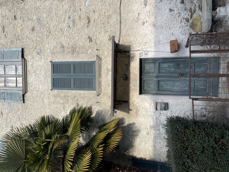 An upward-facing outdoor shot captures the facade of a rustic building, likely in Castione, Italy, under bright sunlight. The wall is made of light-colored, textured plaster or stucco, possibly incorporating visible stones.
On the left, two windows are visible. The upper window has multiple panes and partially open, weathered blue-grey wooden shutters, revealing sheer white lace curtains inside. Below it, a smaller window also features open blue-grey shutters. In the center, there's another window with closed blue-grey louvered shutters.
To the right, a matching blue-grey double-paneled wooden door serves as the entrance. Above the door, a small sign displays the numbers "15" and "401". A small wooden box hangs from chains attached to the wall above and to the right of the door. Below the door, a silver-colored mailbox is affixed to the wall. Further right, a decorative wrought-iron gate is visible.
A prominent palm tree occupies the lower left and central portions of the image, its fronds casting sharp, elongated shadows across the sunlit wall, indicating a clear, sunny day. A dark green bush or hedge is visible in the lower right corner. A dark horizontal beam or shelf structure extends across the facade between the central window/door and the left windows. No people or specific activities are depicted; the scene is static.