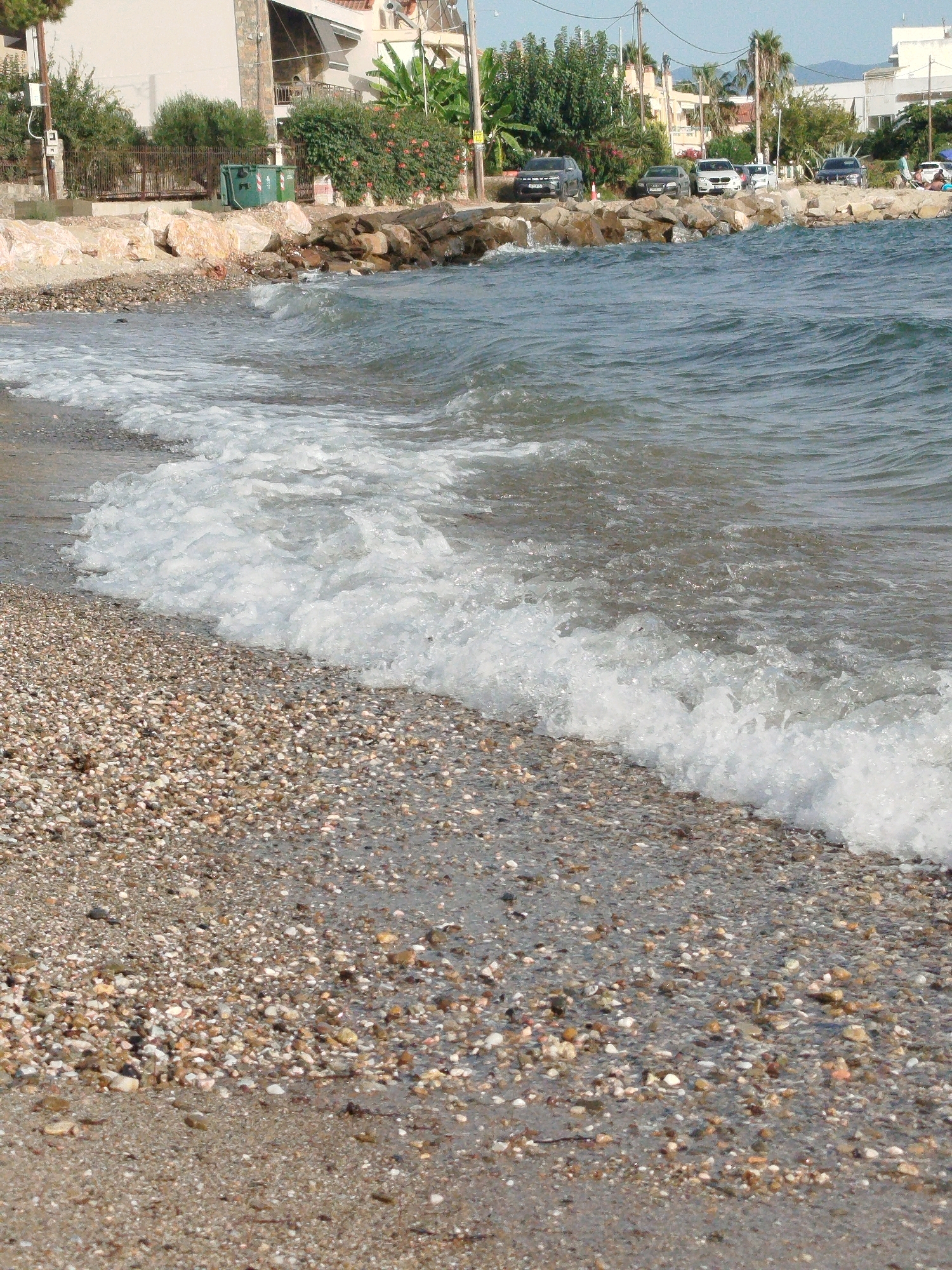The image shows a pebbled beach with the sea gently washing onto the shore. In the background, there are buildings, greenery, and parked cars, suggesting an urban or semi-urban coastal area. The presence of trash bins and a fence indicates some level of infrastructure. The overall scene depicts a typical coastal environment in a developed area.