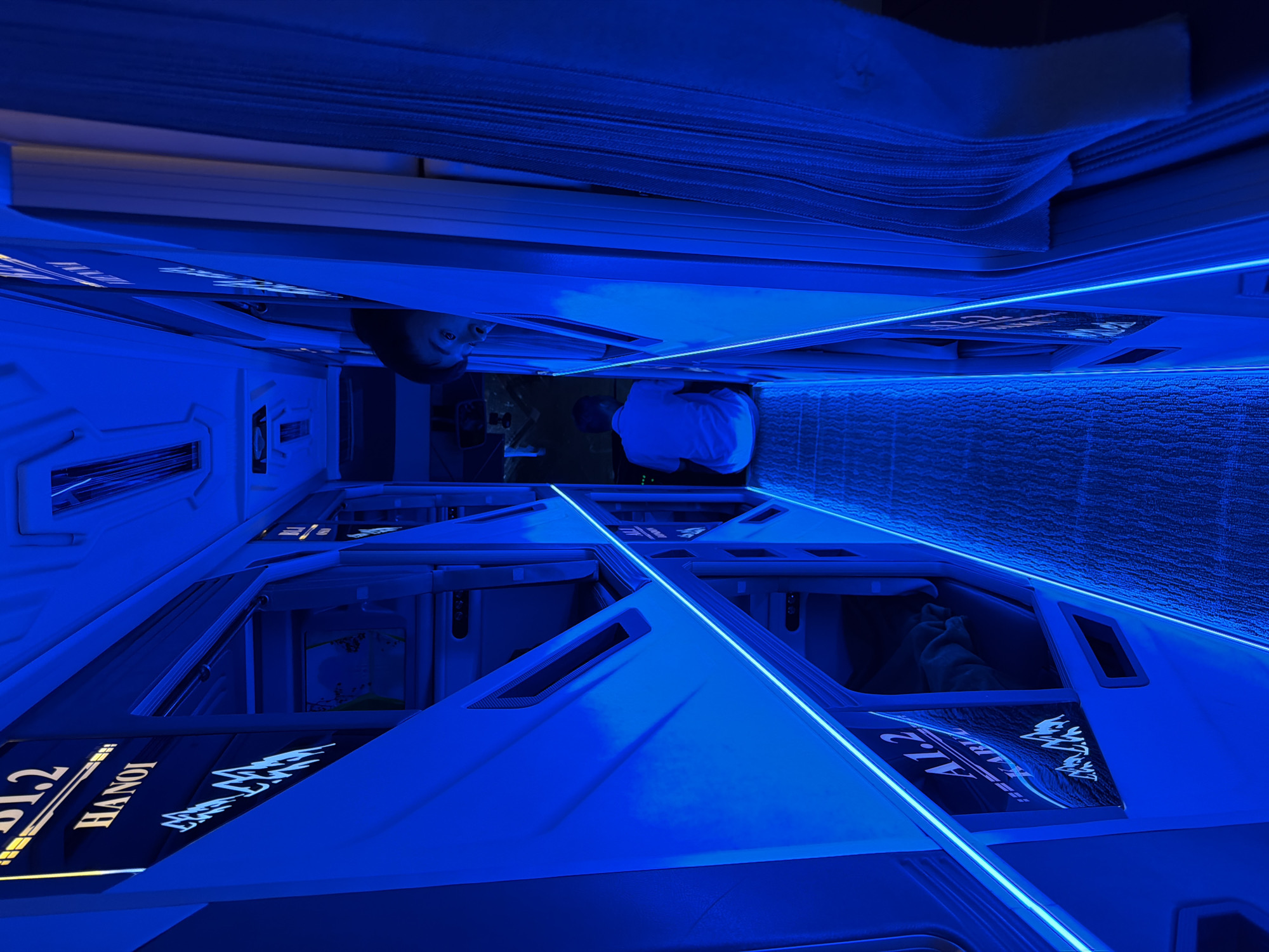 This image captures the interior of a modern capsule hostel or hotel, bathed in a striking electric blue light. The scene depicts a corridor lined with futuristic sleeping pods, viewed from a slightly elevated perspective looking down the length of the hallway.
Two individuals are visible within this blue-hued environment. On the left side of the frame, a person with dark hair is seen peeking out from the opening of a capsule, looking directly towards the viewer with a curious or observant expression. Further down the corridor, on the right, another individual is visible from behind, seated or kneeling inside their open pod, wearing a white shirt. They appear to be engaged in an activity within their private space, possibly settling in or interacting with a device.
The dominant feature of the scene is the sleek, angular design of the capsule beds, accentuated by bright blue LED strip lighting running along their edges and possibly within the ceiling or wall structures. The pods themselves feature smooth, light-colored surfaces with dark accents. One pod prominently displays the text "A1.2 HANOI" along with stylized, intricate script or symbols, suggesting a location in Vietnam. The upper part of the image shows textured material, likely a ceiling or wall covering, also illuminated by the blue light.
The pervasive blue lighting creates a cool, modern, and somewhat futuristic atmosphere, indicating an interior setting where natural light is absent, likely at night. The design suggests a compact yet stylish accommodation experience, common in urban areas like Đà Nẵng, Vietnam.