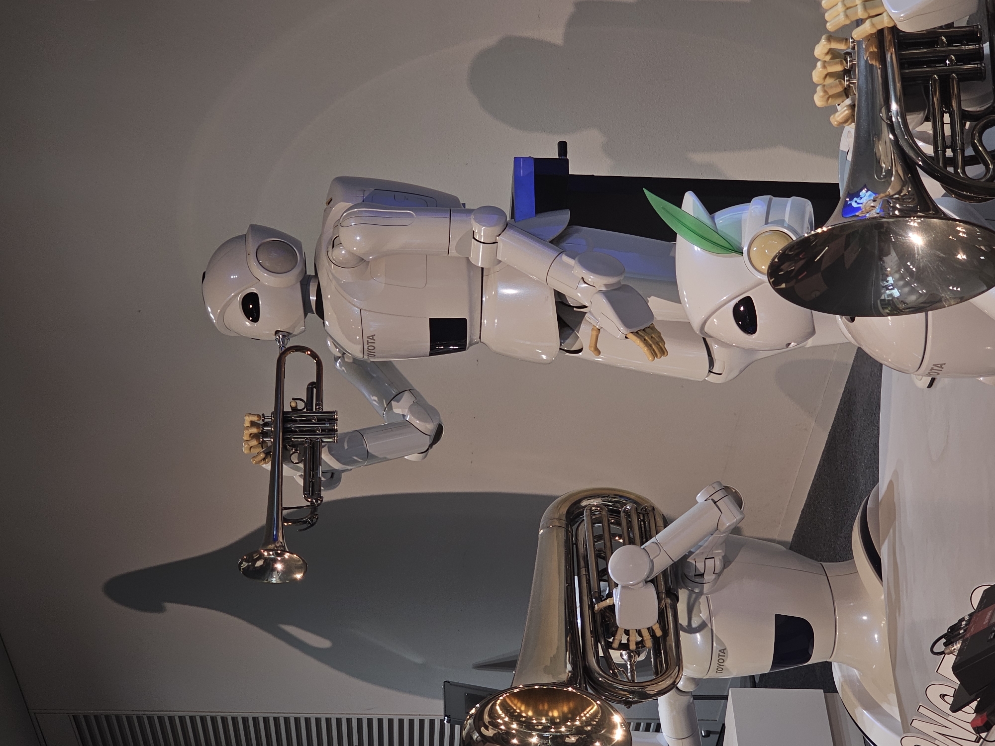 In a museum setting in Nagoya, Japan, three humanoid robots are depicted with musical instruments. The robot in the foreground left holds a tuba, its robotic arms supporting the instrument. A second robot, positioned centrally, plays a trumpet, its hands on the valves and its mouth near the mouthpiece. A third robot, partially visible in the background right, also appears to be holding a musical instrument. The robots are white and sleek, with visible joints and articulated hands. They are positioned on a floor that appears to be carpeted, with a plain gray wall as the backdrop. The lighting casts shadows on the wall, adding depth to the scene. The robots are branded with the "TOYOTA" logo.
