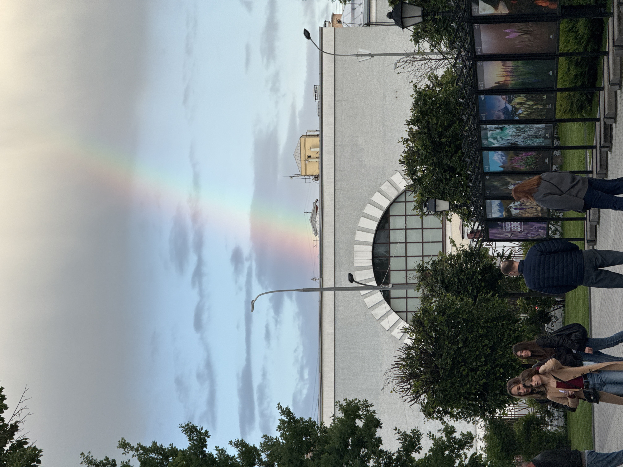 The image depicts a street scene in Moscow, Russia, under a cloudy sky with a distinct rainbow stretching across the left side of the frame. The rainbow's base appears to touch the ground.
To the right of the rainbow is a light gray building, likely a museum or cultural institution, with a large, semi-circular window. A street lamp is positioned in front of the building.
Further to the right, a black, wrought-iron structure displays a series of framed images, possibly an outdoor photography exhibition. Several people are present, including a woman with red hair looking at the exhibition, a man in a dark jacket walking nearby, and a group of three young women in casual attire walking along the sidewalk.
The foreground is filled with trees and bushes, framing the scene. The overall impression is of a public space where people are engaging with art and enjoying a rare natural phenomenon.