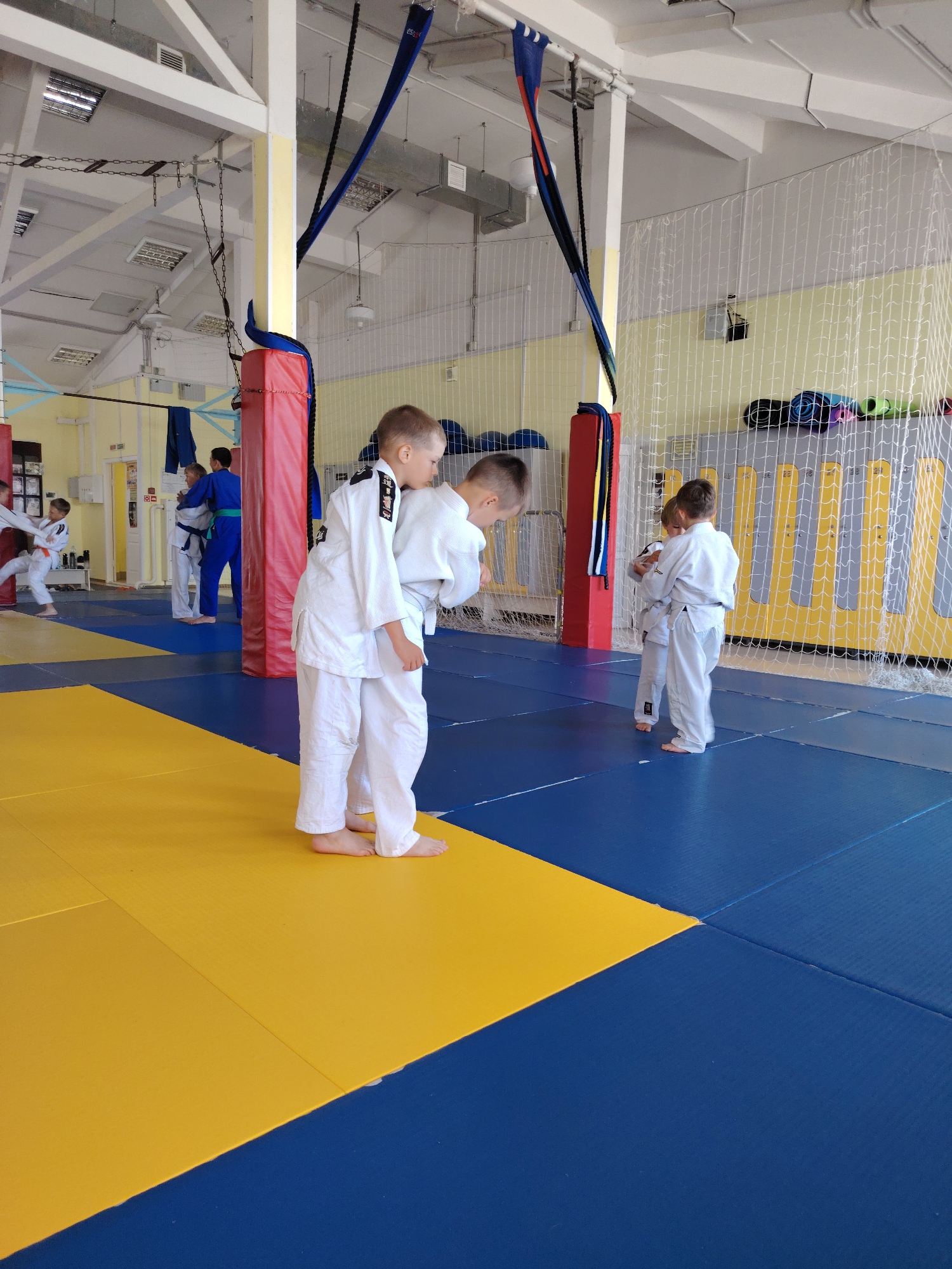 The image depicts an indoor martial arts training facility, likely a judo or wrestling dojo, located in Chaikovsky, Russia. The floor is covered with a blue and yellow mat system. In the foreground, two young boys, dressed in white judogi (martial arts uniforms), are engaged in practice. The boy on the left, facing away from the camera, is standing close to the boy on the right, who is also facing away and appears to be demonstrating or receiving instruction.
In the mid-ground, another young boy in a white judogi is practicing with an instructor or another student in a blue judogi with a green belt. Further in the background, more individuals in judogi are visible, suggesting a class or training session is in progress.
The environment includes typical dojo features such as padded posts, a net enclosure (possibly for safety or to demarcate an area), and overhead lighting and ventilation systems. The overall impression is one of a structured and active training environment.