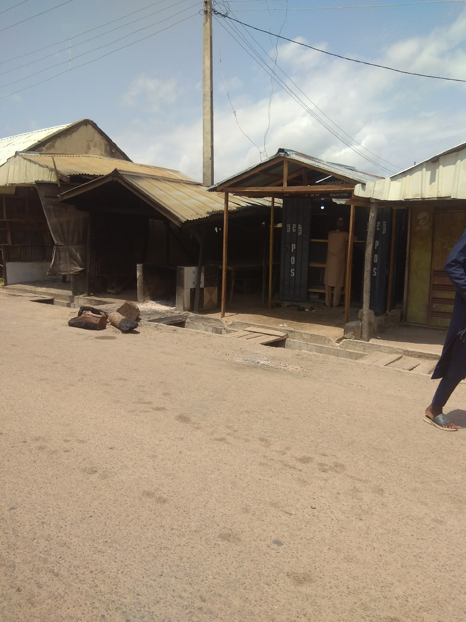 The image depicts a street scene in Jalingo, Nigeria, under a bright, cloudy sky, suggesting daytime. The foreground is dominated by a paved road with some scattered debris, including several pieces of wood that appear to be logs.
In the midground, several market stalls or shops line the street. These structures are characterized by their corrugated metal roofs and open fronts. One stall, partially visible on the right, has a yellow facade. Another stall in the center features a covered wooden frame and appears to be constructed from repurposed shipping containers, with the text "POS" visible vertically on the blue panels of the containers. A person in a light brown garment stands inside this stall, facing away from the camera. Another stall to the left has a more rustic appearance with a dark, corrugated metal exterior and a fabric awning.
The background shows more of the market structures and utility poles with overhead electrical wires, common in urban Nigerian settings. On the far right, a person in a dark blue garment and sandals is walking away from the camera. The overall impression is of a busy, informal marketplace, typical of many towns in Nigeria. The weather appears warm, as indicated by the bright sky and the clothing of the individuals.