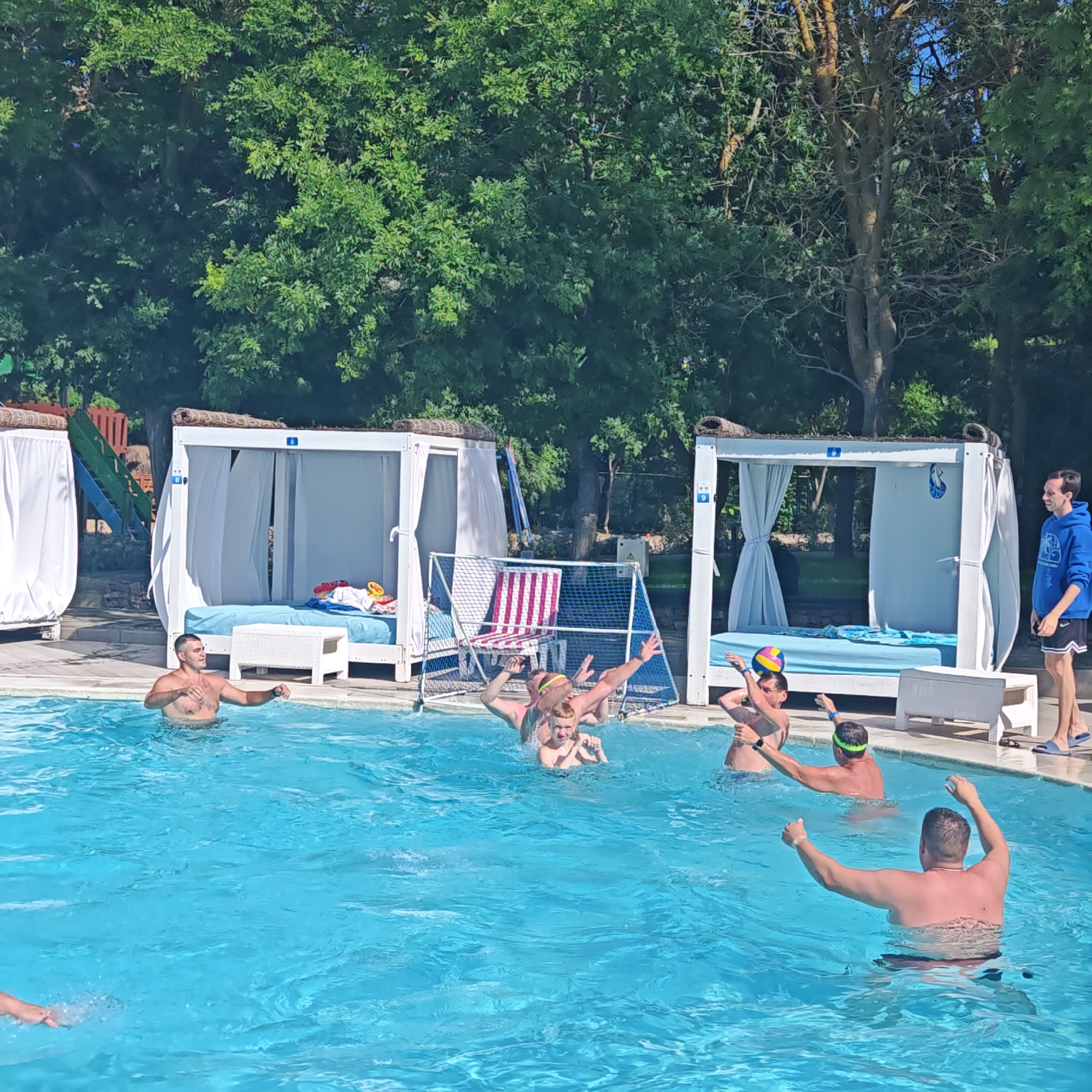 In a bright, sunny outdoor setting, a group of people are enjoying a day at a swimming pool. The pool is a vibrant blue, with clear water. In the background, there are several white cabanas with draped curtains, some of which have blue cushions on them, suggesting a resort or relaxation area. A water polo net is set up in the pool, indicating a game or recreational activity is in progress.
Several individuals are actively engaged in the pool. One man, shirtless, stands in the water with his arms outstretched, appearing to be playing or interacting. Further in the pool, a group of people are in motion, some with arms raised, possibly in the midst of a water polo game. One person is wearing a green swimming cap or headband. Another person in the pool is tossing a multi-colored ball. To the right of the pool, a man in a blue hoodie and black shorts stands on the edge, watching. His clothing suggests a casual, possibly warm weather, but not extremely hot, atmosphere.
The overall scene depicts a fun, active day outdoors. The presence of the cabanas and pool suggests a leisure facility. The sunlight and clear blue sky indicate good weather. The emotions conveyed are those of enjoyment and activity. No visible text is apparent on the people or objects in the image, apart from what might be small logos or labels on the cabanas that are not legible. The provided location context of "городское поселение Щелкино, Ukraine" suggests this is a public or recreational space in that region.