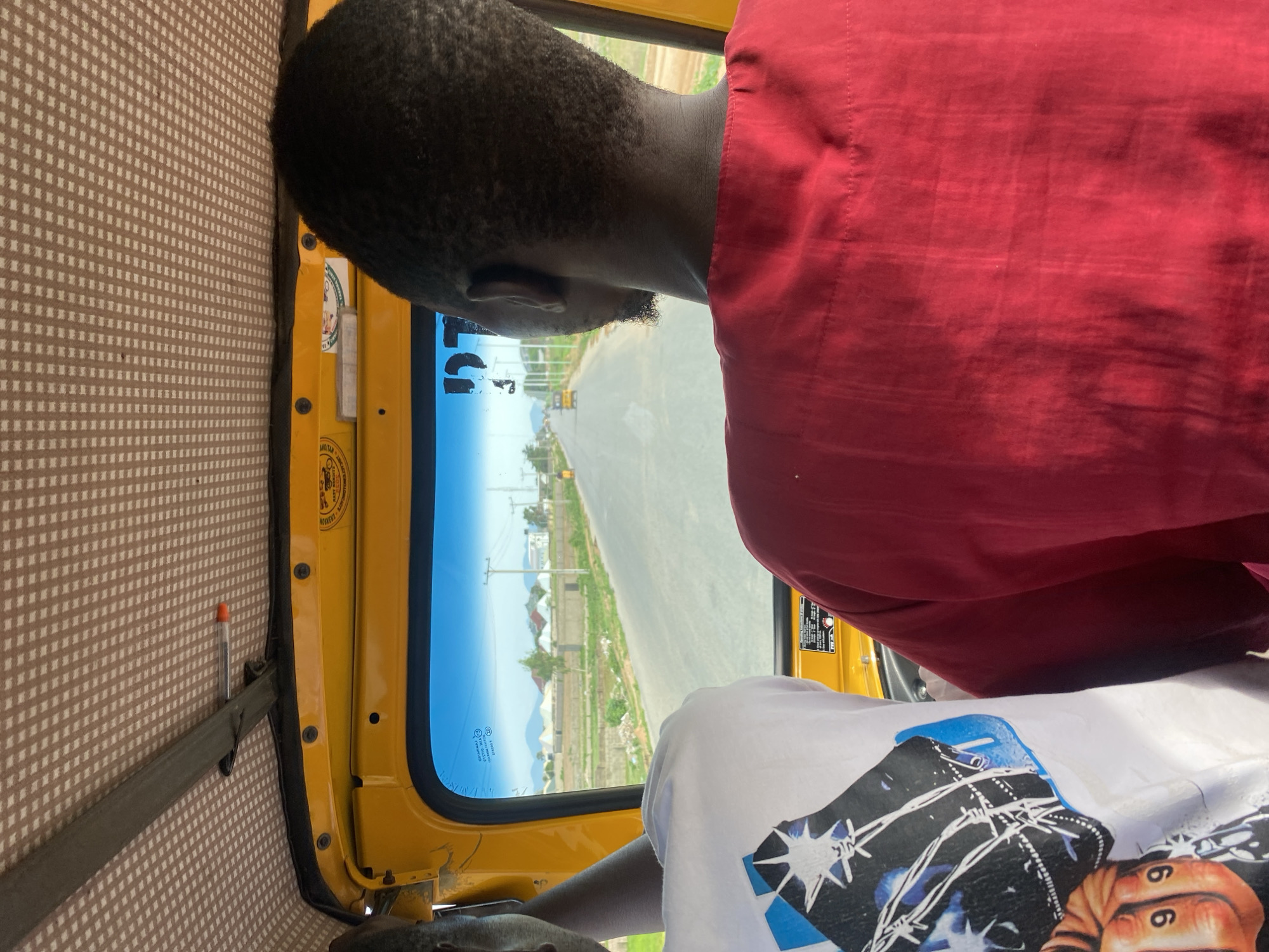 This media file captures a view from inside a yellow vehicle, likely a tuk-tuk or similar auto-rickshaw, looking out through the passenger side window. The primary subjects are two people. One person, seen from behind, wears a vibrant red garment and has short, dark, textured hair. Their head is turned slightly, suggesting they are looking out the window or interacting with someone in the vehicle. Another person, partially visible in the foreground, wears a white t-shirt with a graphic design featuring abstract shapes, barbed wire, and the number "66".
The scene outside the window depicts a paved road stretching into the distance, with a rural or semi-urban landscape on either side. Green vegetation, scattered buildings with distinct roofs, and utility poles are visible. The sky is bright and appears clear, suggesting it is daytime. The light indicates a sunny day, possibly late morning or afternoon.
Inside the vehicle, the brown and white patterned interior lining is visible on the left. A dark strap is attached to the wall, with a pen clipped to it. The yellow frame of the vehicle's body and window are prominent. There are some stickers or labels on the yellow frame near the window's edge, one of which appears to have some text and an image, possibly indicating branding or regulations.
The overall impression is of a journey in progress on a warm, sunny day in Jalingo, Nigeria. The perspective suggests the image was taken from a passenger's viewpoint, looking outwards. There are no strong emotional cues evident, but the activity appears to be casual travel.