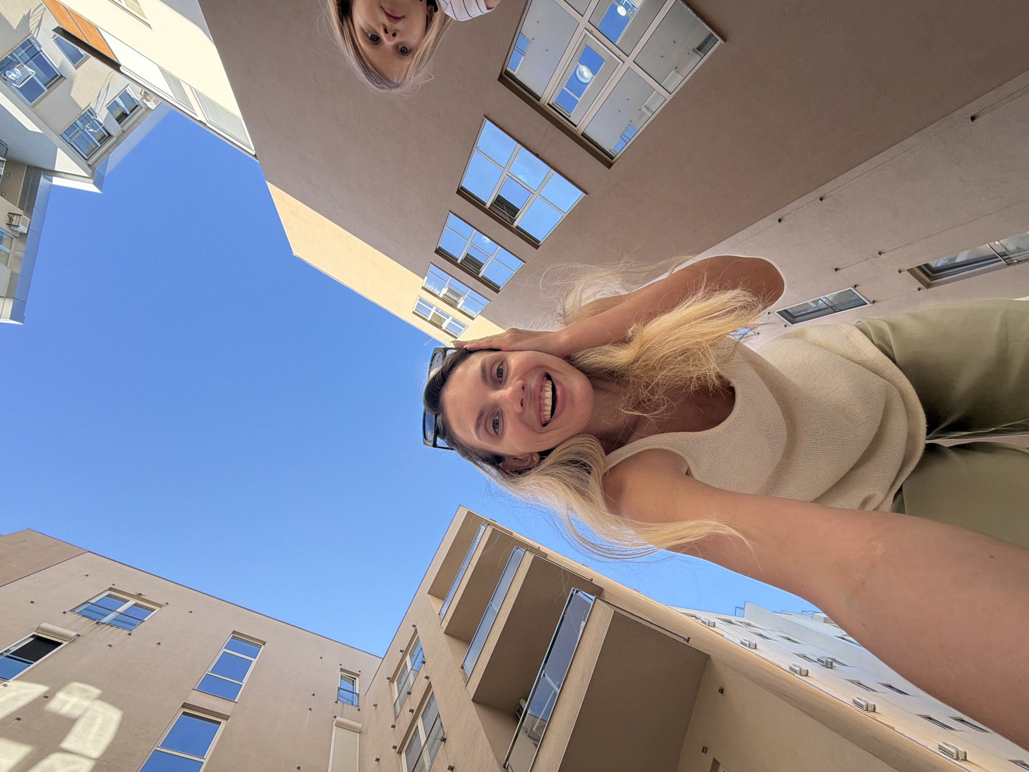 A low-angle, outdoor, wide shot captures a woman in the foreground, smiling and looking upwards, with a child to her right, also looking upwards. The woman has blonde hair and is wearing a light-colored sleeveless top and green pants. The child has blonde hair and is wearing a striped shirt. They are situated between tall, modern buildings with visible balconies and numerous windows. The sky is clear and blue. The buildings appear to be residential or hotel apartments, constructed with light-colored facades and glass balconies. The architectural style suggests a contemporary urban setting, possibly in the city of Budva, Montenegro, known for its modern coastal architecture. The image is taken from a perspective looking up into the sky, emphasizing the height of the buildings and the overhead view.