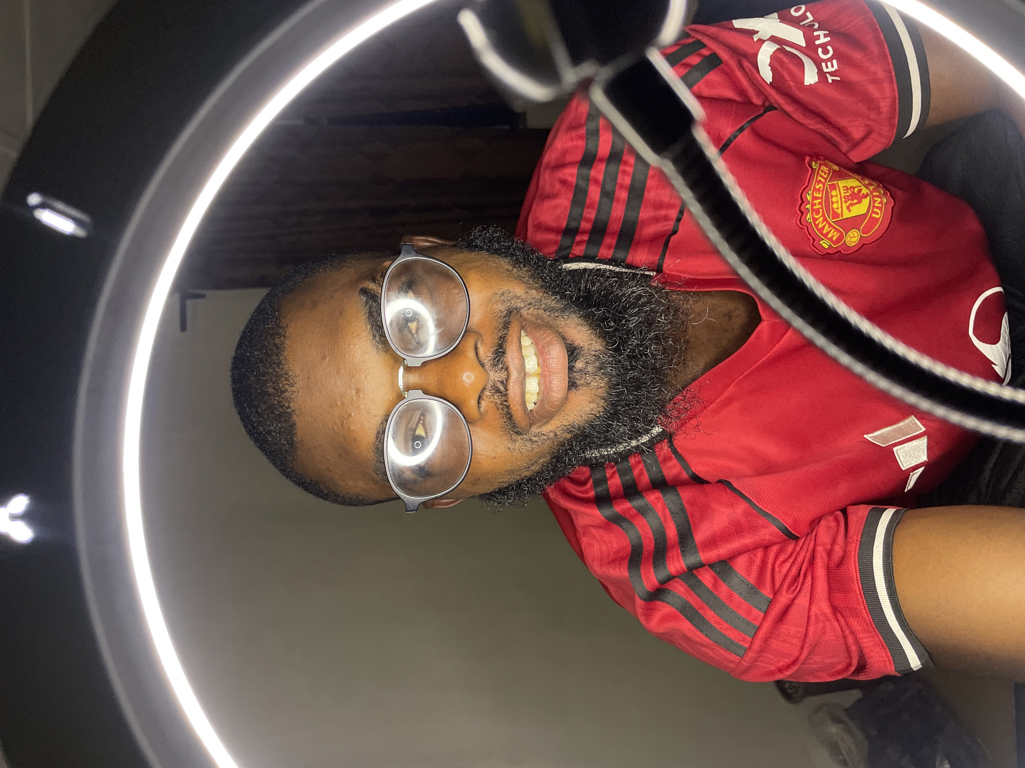 A medium shot captures a Black man from the chest up, wearing a red Manchester United jersey with black stripes. He has a beard and is wearing round, silver-rimmed glasses. The man is smiling, revealing his teeth, and is positioned in front of a ring light that creates a bright halo effect around his head. The background is a plain, dark wall, suggesting an indoor setting. The man appears to be in his home or a studio, possibly preparing for a video recording or photo shoot. The scene is a personal portrait, not indicative of any specific urban landscape or public space in Barakin Lerawi, Nigeria, or elsewhere.