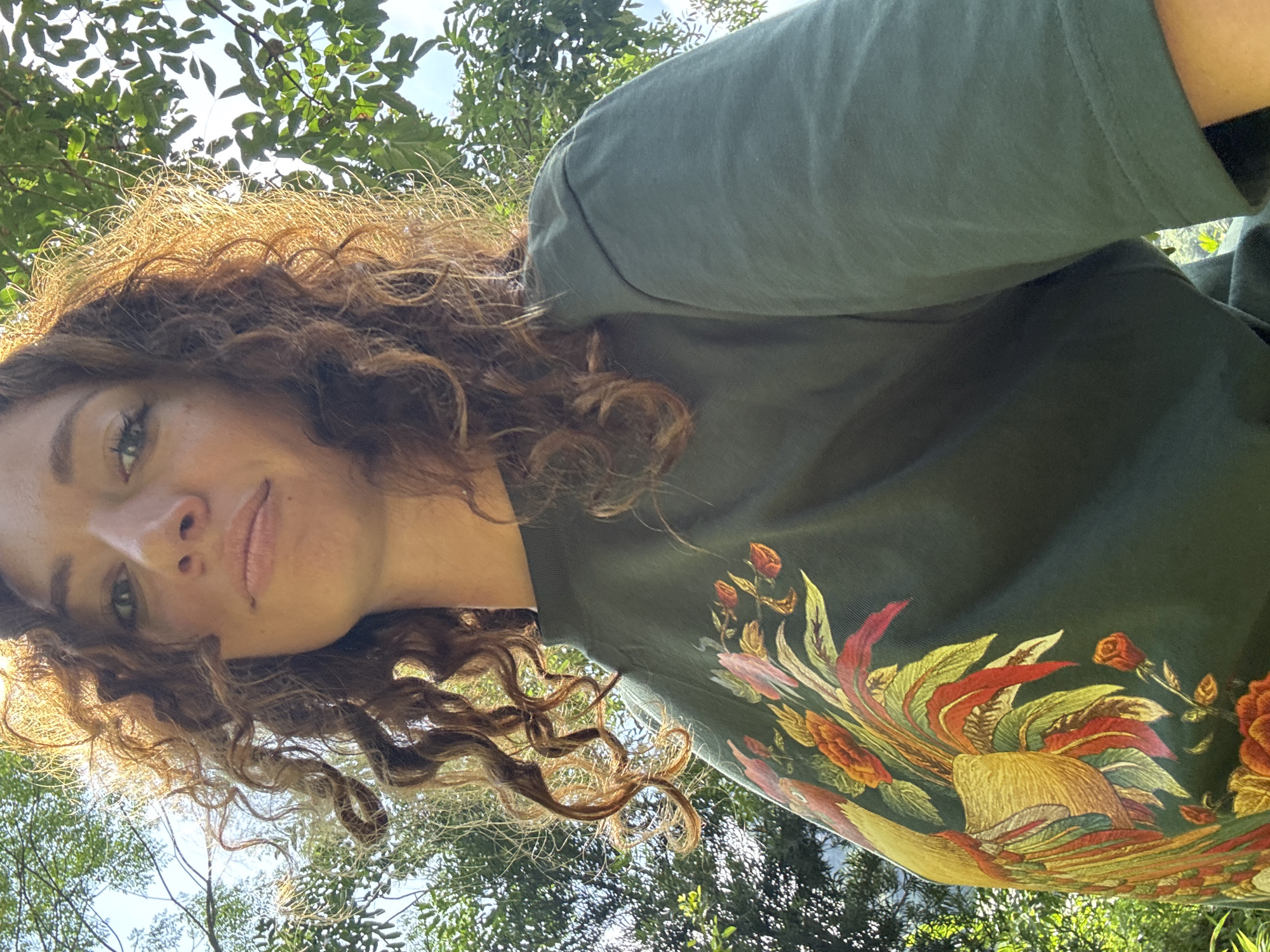 A young woman with curly, auburn hair is looking up at the camera, her face partially obscured by her hair. She is wearing a dark green t-shirt with a large, colorful embroidered design of a bird and flowers on the front. The background consists of lush green foliage and a bright sky, suggesting an outdoor setting. The lighting indicates it is daytime, possibly late morning or early afternoon. The image appears to be a selfie taken outdoors.