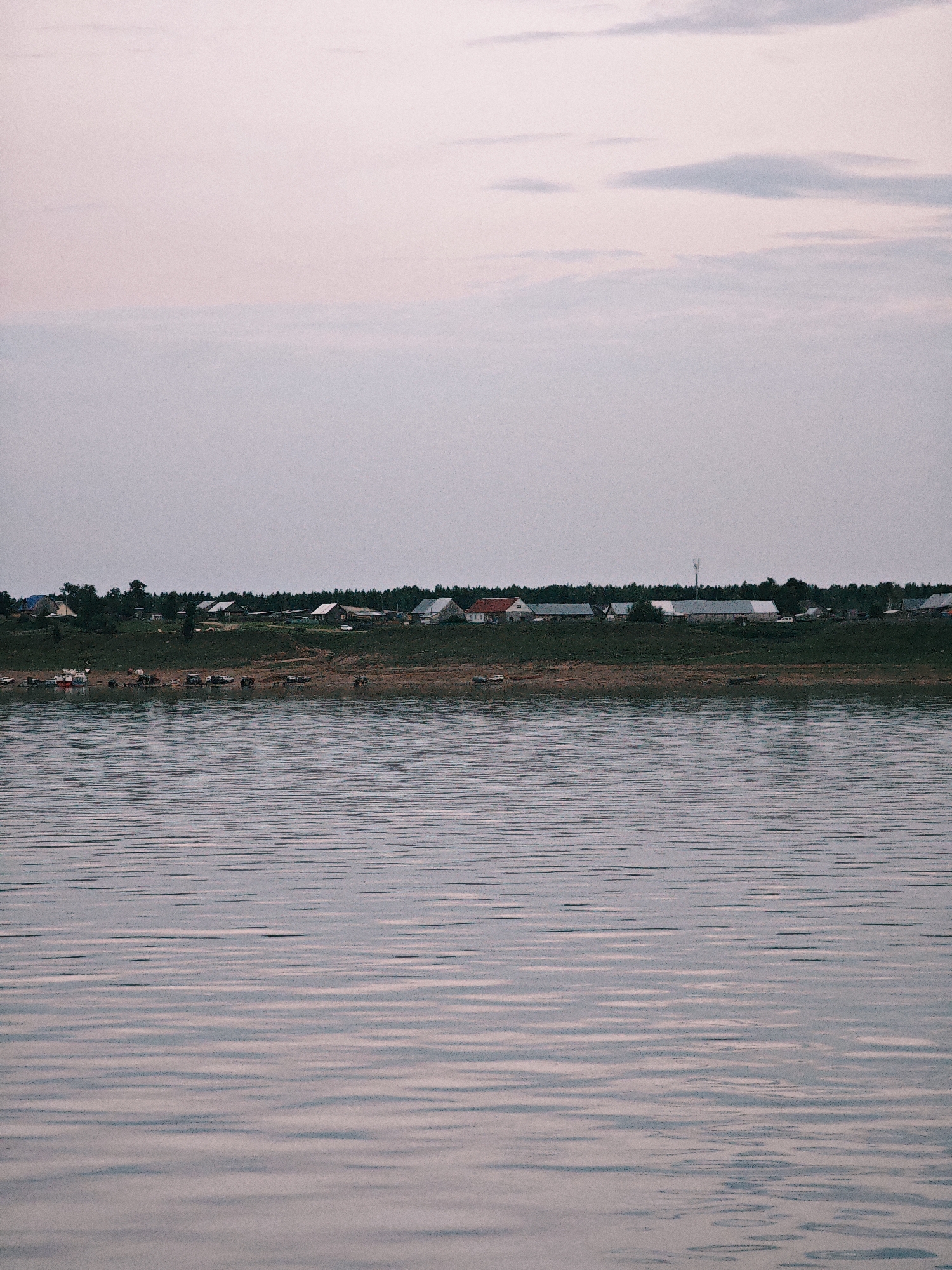 The image depicts a serene landscape with a body of water in the foreground and a village nestled on a hill in the background. The water's surface is rippled, reflecting the muted, pastel hues of the sky. The sky itself is a soft gradient of pink and pale blue, with a few wispy clouds.
The village appears to be a rural settlement, characterized by a cluster of houses with pitched roofs, interspersed with trees and some open grassy areas. Some of the houses have visible chimneys. A line of dense trees forms a backdrop to the village, suggesting a forested area beyond. On the grassy slope leading down to the water, there are several vehicles, including what appear to be cars and possibly some agricultural machinery. A small boat is also visible near the water's edge. The overall impression is one of a quiet, undeveloped location.
The time of day appears to be either early morning or late evening, given the soft, diffused light and the pastel colors of the sky. The weather seems calm, with no strong winds suggested by the water's surface. The location is identified as Nikulino, Russia. The scene evokes a sense of peace and tranquility, with the natural elements of water and sky dominating the composition. There is no visible text in the image.