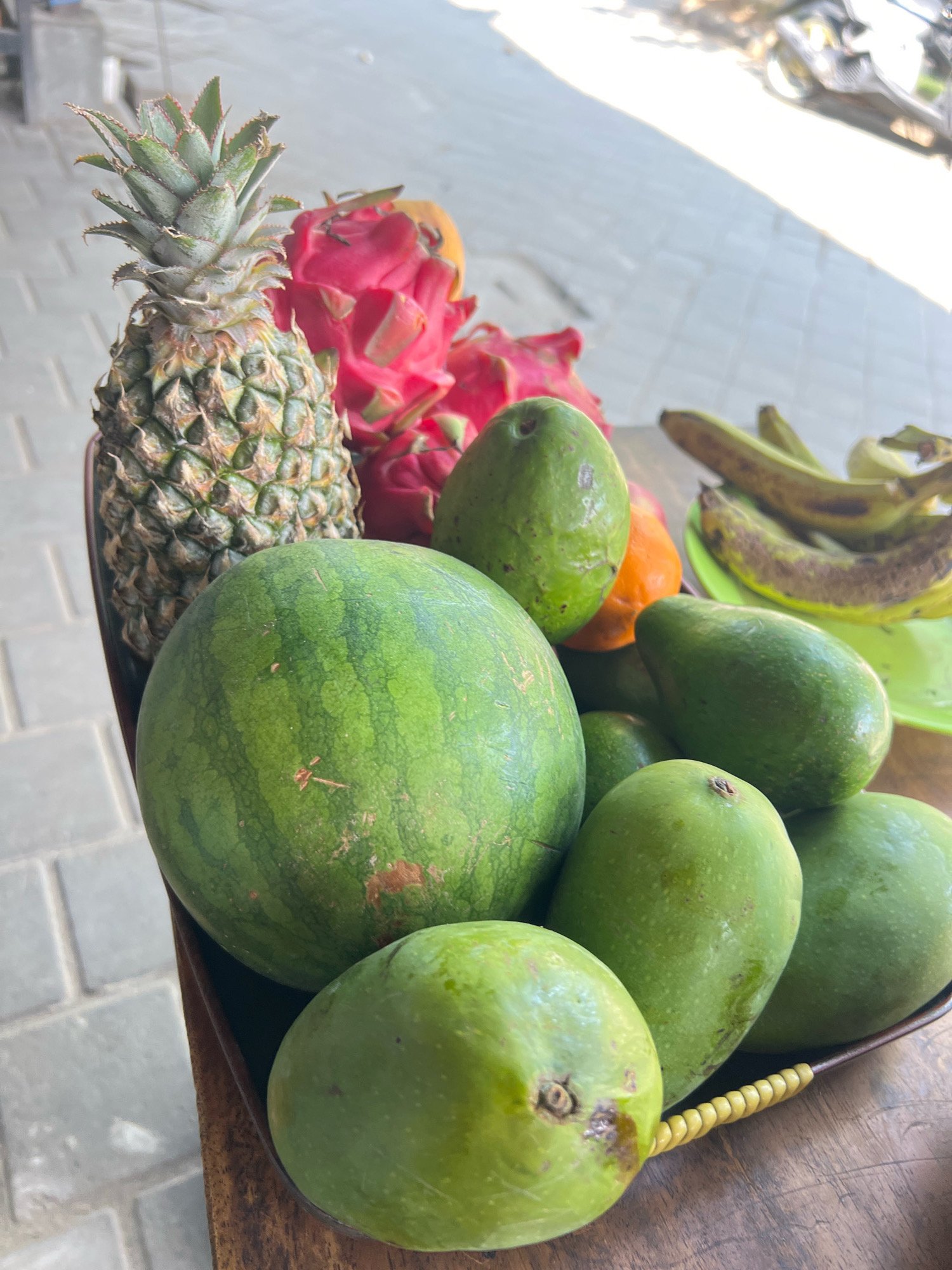 In Kuta, Indonesia, a close-up shot captures a vibrant assortment of fresh fruits arranged on a rustic wooden table, likely outdoors given the tiled ground and dappled sunlight. Dominating the foreground is a large, green watermelon with subtle stripes, followed by several ripe, green mangoes. Behind the watermelon, a pineapple stands tall, its spiky crown reaching upwards, juxtaposed with bright pink dragon fruits. A few green avocados are scattered among the fruits, and a single orange fruit, possibly a papaya or cantaloupe, peeks out from behind them. In the background, a collection of overripe bananas rests on a green plate, hinting at a potential sale or distribution. The scene is devoid of human presence, with the focus solely on the bountiful display of produce, bathed in the natural light.