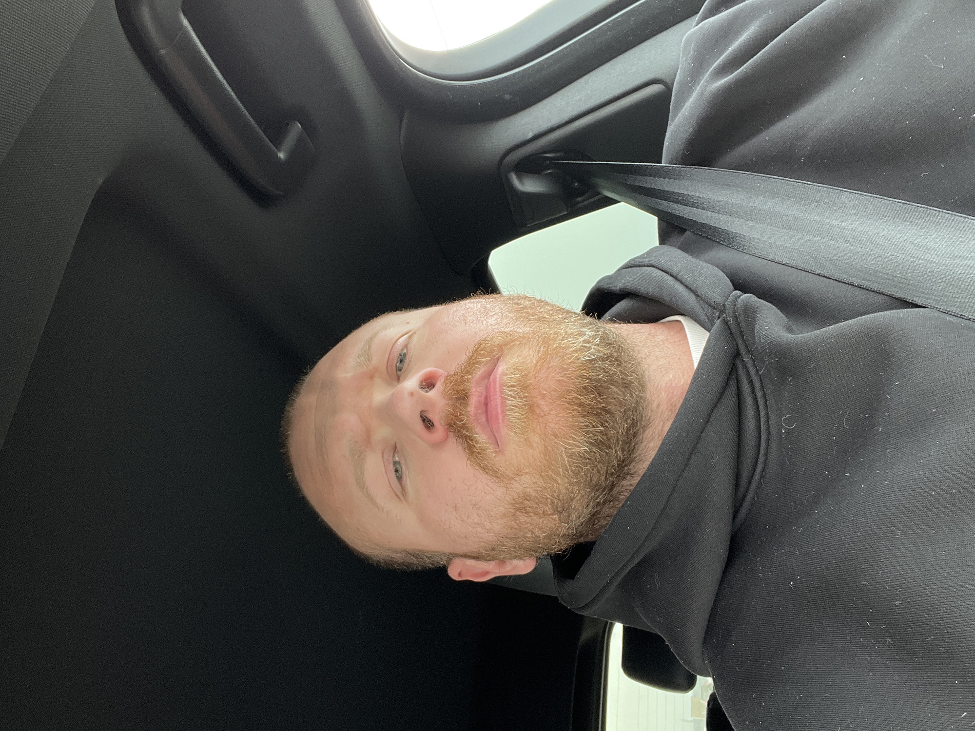 An adult male with fair skin, light-colored eyes, and a reddish-brown full beard and mustache is depicted. He has short hair or is balding on the crown. A small dark mark is present on the bridge of his nose. He wears a black hooded sweatshirt over a white undergarment, with visible lint on the sweatshirt. He is seated, looking directly at the camera, with a black seatbelt strap secured across his chest from the upper right to the lower left. The setting is the interior of a vehicle, characterized by a dark-colored textured headliner and interior trim. A dark grab handle is visible on the vehicle's ceiling to the upper right. The exterior environment, visible through the vehicle's windows, appears as a bright, undifferentiated light source. The image provides no details to ascertain a specific geographical location, including the city of Chaikovsky, Russia.