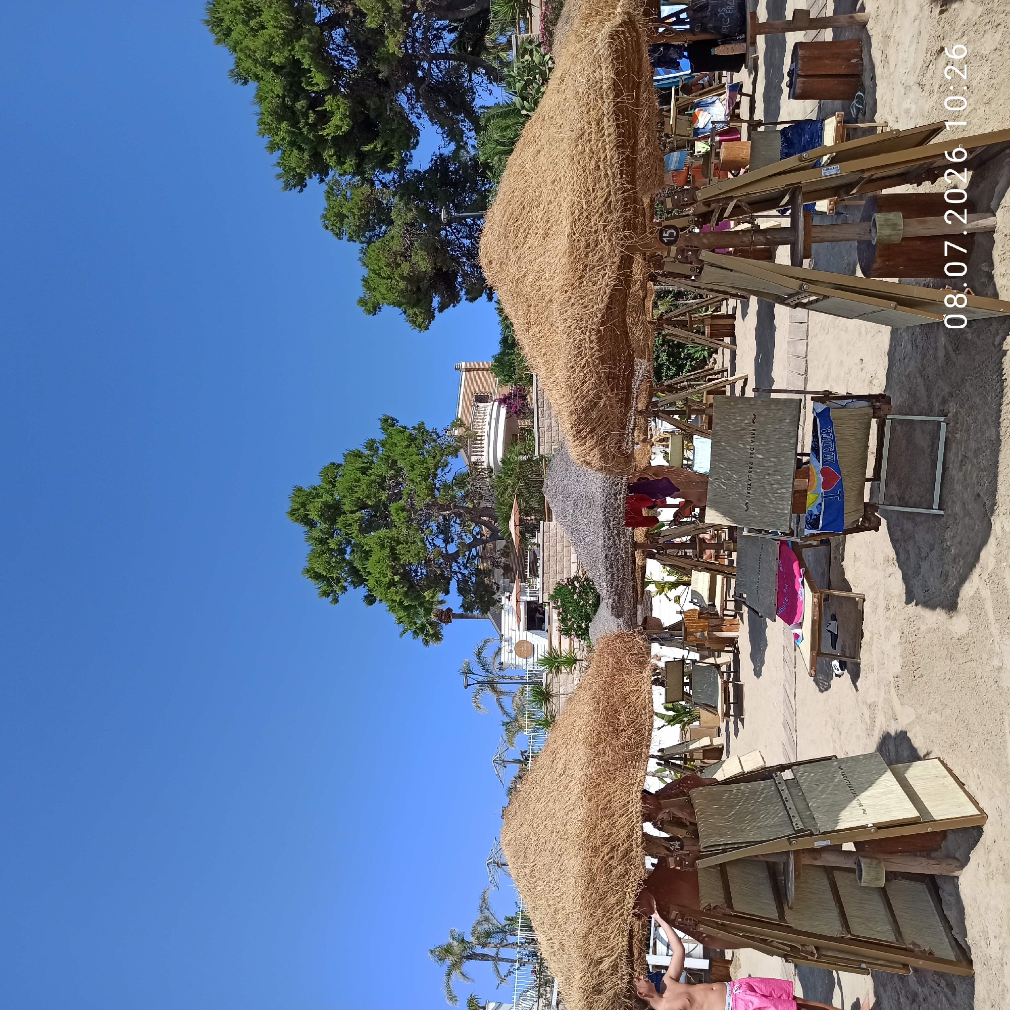 This is a wide, low-angle shot of a sandy beach in Trani, Italy, on a clear, sunny day with a bright blue sky. In the foreground, two thatched umbrella structures with wooden supports are prominent. Underneath these umbrellas are rows of wooden lounge chairs and small tables, some with towels and bags on them. Several chairs are folded and leaning against the supports.
A young person, wearing pink shorts, stands to the left, facing away from the camera and towards the umbrellas, with their left arm resting on one of the structures. In the background, a white building with arched windows and a balcony is visible, surrounded by lush green trees, including palm trees. Some other beach amenities, such as a small white building with signage and more thatched umbrellas, are also present. The sand appears dry and well-trodden. A date and time stamp, "08.07.2026 10:26," is overlaid on the lower right portion of the image.