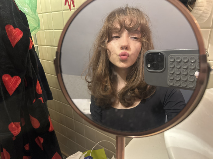 The image depicts a mirror selfie taken by a single individual in what appears to be a private room, likely a bathroom, within the city of Unknown, Unknown. A young female subject with light brown, wavy hair and bangs, displaying a septum piercing, is reflected in a round, copper-framed vanity mirror. She wears a black long-sleeved top and has her lips pursed in a pout. She holds a dark gray smartphone, featuring a textured gray "pop-it" style case and visible triple camera lenses, in her right hand (viewer's perspective) to capture the image. A white painted thumbnail is visible on the hand holding the phone.
To the left of the mirror, a black garment patterned with large red hearts is draped or hanging. Above it, a translucent green plastic bag is visible. The wall behind these items is tiled with light yellow/pale green square tiles. A portion of a white sink is visible in the bottom right corner, partially obscured by the mirror's stand. Other obscured objects, including a red ribbon and additional items on a surface, are present in the background.
