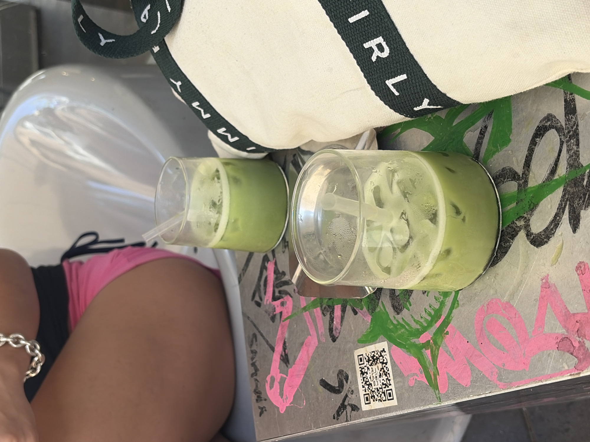 The image captures a close-up view of two glasses of iced green drinks, likely matcha lattes, placed on a metallic table with graffiti-style markings in pink, black, and green. A white canvas bag with dark green trim featuring the word "GIRLY" in white lettering is partially visible on the right. In the background, a person's legs are visible, wearing bright pink shorts and a silver bracelet. The setting appears to be an outdoor cafe or a casual outdoor seating area, possibly in Tel Aviv, Israel, given the graffiti art style commonly found in urban areas there. The drinks are served in clear glasses with straws. The overall atmosphere suggests a relaxed, informal setting.