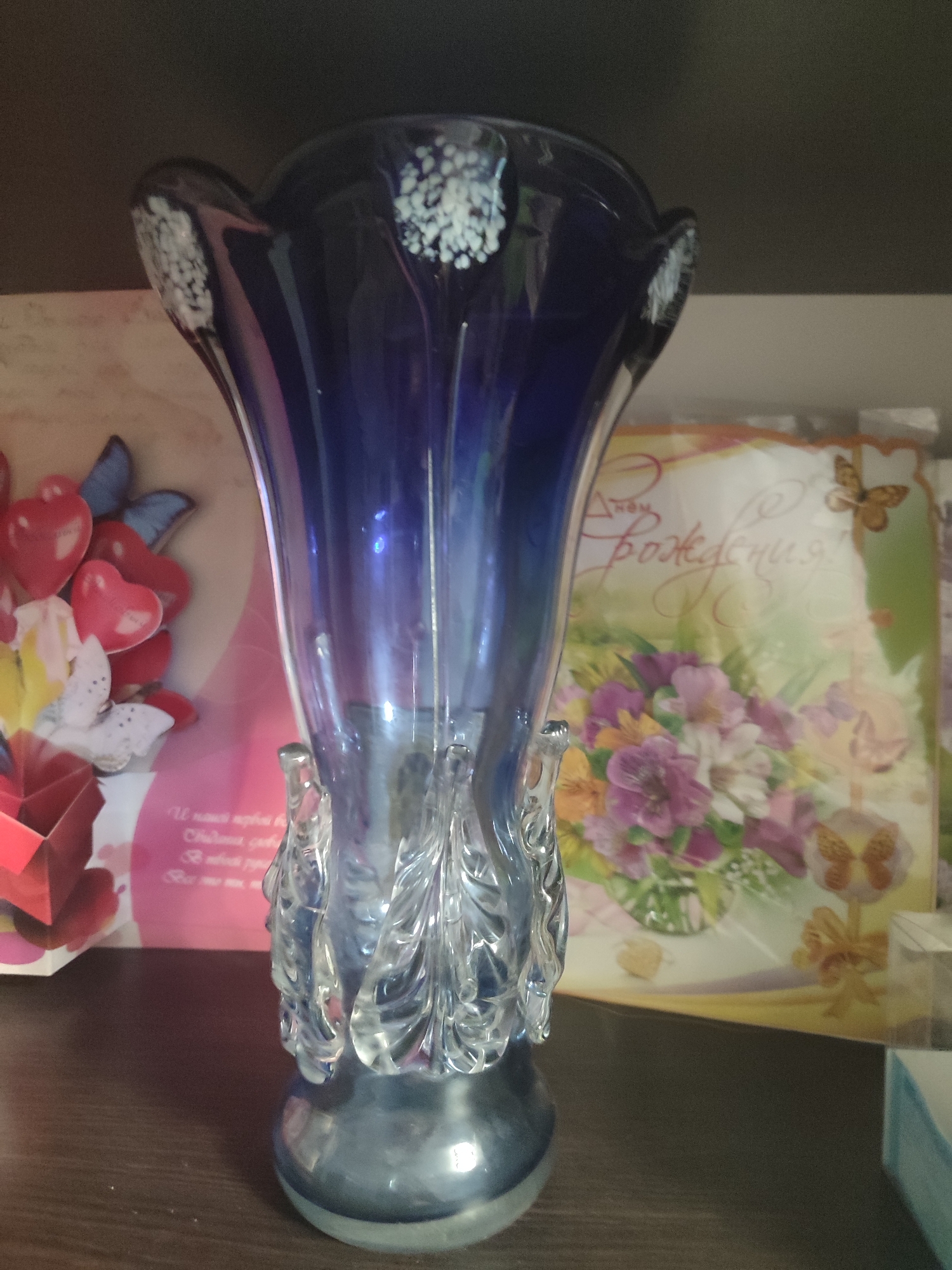 The image shows a glass vase with a dark blue top and a clear, textured base, positioned on a wooden shelf. The vase is in the foreground and occupies the majority of the frame. Behind the vase, to the left, is a greeting card featuring red hearts and white butterflies. To the right of the vase is another greeting card with a floral design, pink and purple flowers, and butterflies, along with Russian text that appears to say "С Днём Рождения!" (Happy Birthday!). The background consists of shelves and a dark wall. There are no people or animals present in the image, and no specific actions or interactions are depicted. The scene appears to be an indoor setting, possibly a home or a shop, in Mykolaiv, Ukraine.