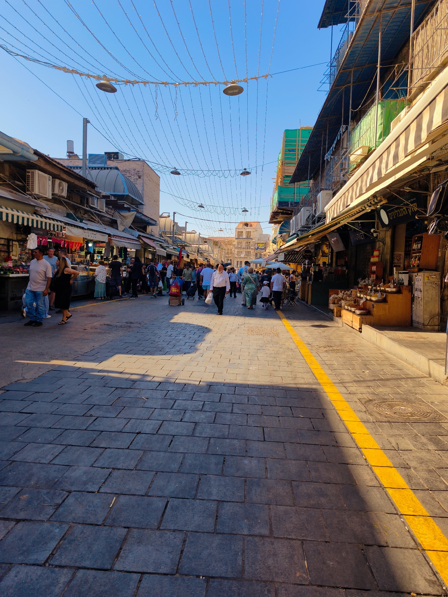 A vertical, eye-level, medium shot captures a bustling street market in Jerusalem, Israel, under a clear, bright blue sky. Strings of decorative lights crisscross the scene overhead. On the left, market stalls with striped awnings display various goods, and shoppers browse. A man in a white shirt and blue jeans stands near a stall. On the right, more market stalls line the street, featuring awnings and displays of goods. People walk along the paved street, which is marked with a bright yellow line. A man in a white shirt and dark pants walks toward the foreground on the right. The paving stones are dark gray. The sun casts long shadows across the street.