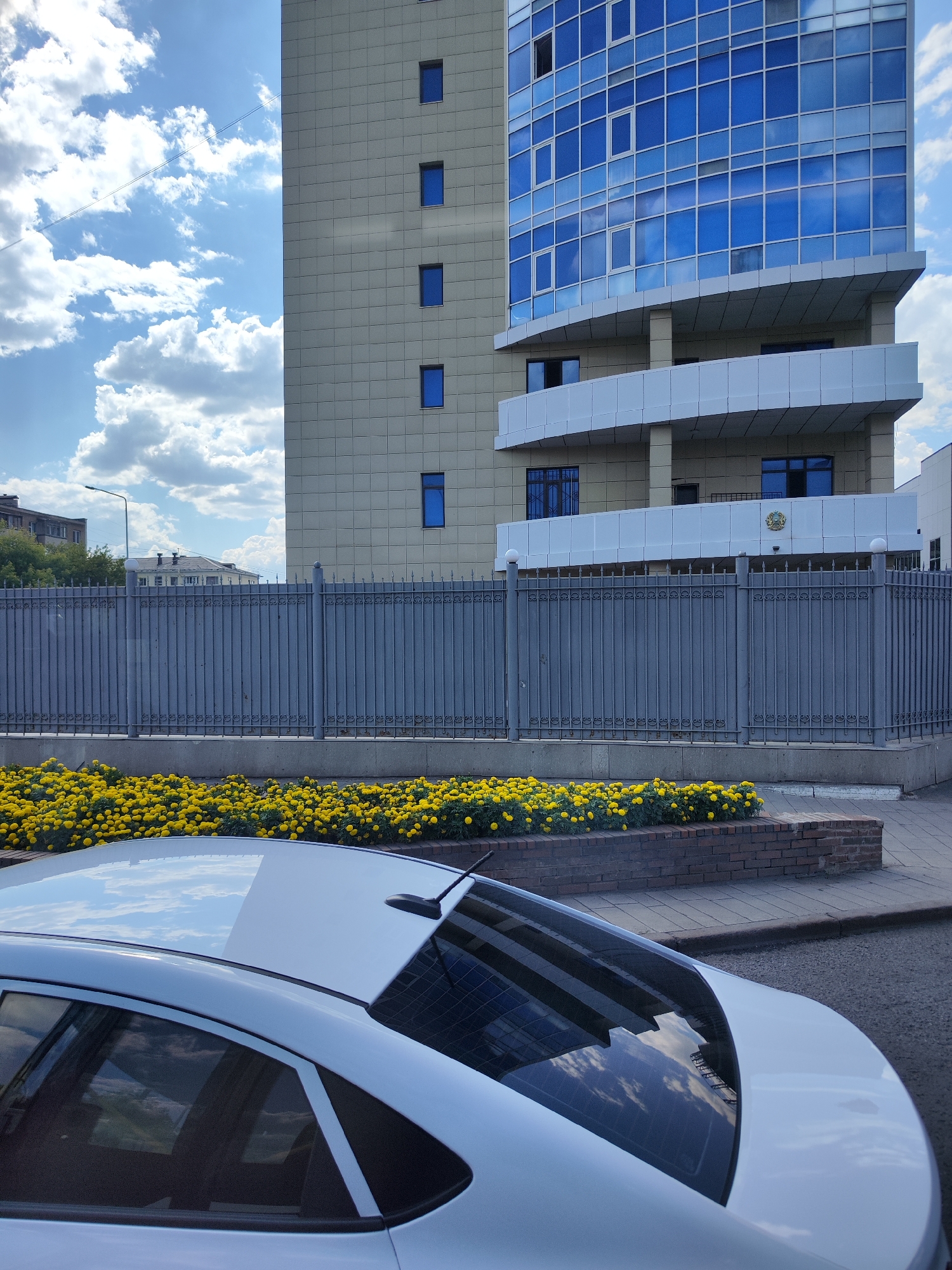 A white sedan is parked on an asphalt street in front of a large, modern building with a beige facade and blue-tinted windows. A grey metal fence with decorative elements separates the street from a flower bed filled with yellow marigolds. The building has a prominent balcony area with a white railing and a coat of arms displayed above the entrance. The sky is blue with white, fluffy clouds, and a thin contrail is visible. The scene appears to be in Astana, Kazakhstan, given the presence of the coat of arms, which is the national emblem of Kazakhstan. The image captures a typical urban streetscape with a focus on architectural elements and landscaping. There are no people visible in the frame.