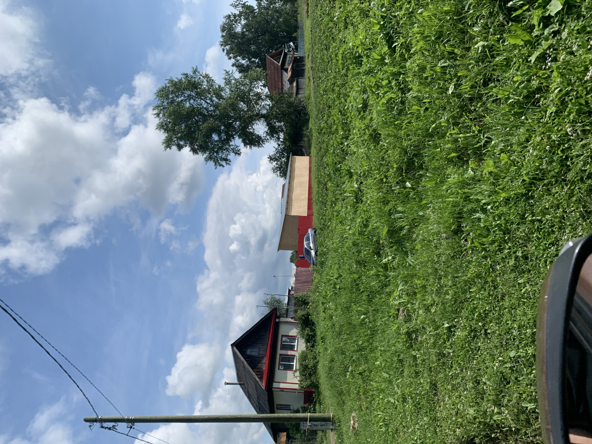 The image shows a rural scene in the city of Barsai, Russia, on a sunny day with scattered clouds. The foreground is dominated by a lush, green meadow. A utility pole with wires stands on the left side of the image. Behind the meadow, there is a small, light-colored house with a red roof and trim, and several red-striped fences. A silver car is parked near the fence. Further in the background, there are several trees and other buildings, including a long, light-colored structure with a red roof. The overall impression is one of a tranquil, semi-rural environment.