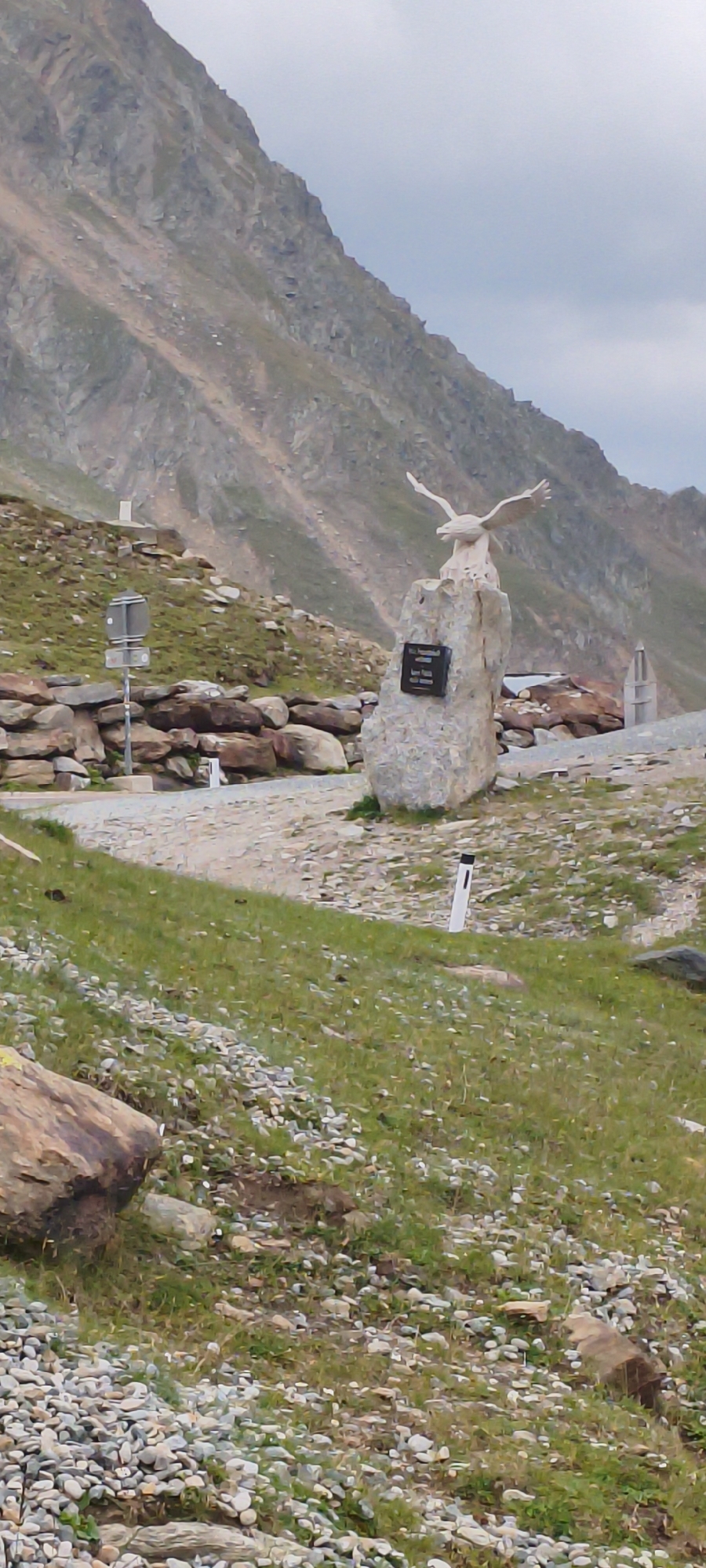 An eagle sculpture perches atop a rock monument located in the mountainous terrain surrounding Sölden, Austria. The sculpture depicts a white eagle with outstretched wings. The monument itself is a rough-hewn stone structure with a dark plaque affixed to its front. In the background, a steep, rocky mountain slope rises, partly covered with sparse vegetation. A paved pathway or road curves through the scene, bordered by a stone wall constructed from numerous rocks. Several white marker posts with black tops are visible along the pathway and on the grassy slopes. The ground is covered with grass and loose stones. The sky is overcast with clouds.