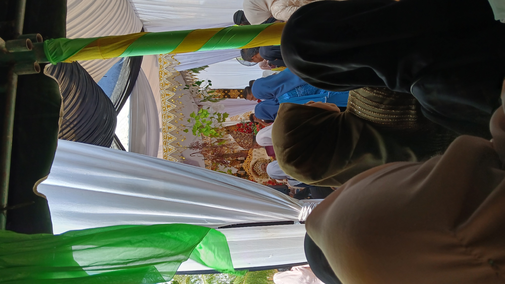 Captured from a low angle inside what appears to be a tent-like structure, the scene focuses on an event occurring within, possibly a wedding. Observers are gathered around. The foreground is dominated by the backs of individuals, their clothing ranging in colors. A central backdrop features a golden-trimmed curtain, and green foliage is placed in front. Hints of architecture and open space are visible through the tent's structure, suggesting an outdoor setting. The location is Pulo, Indonesia.