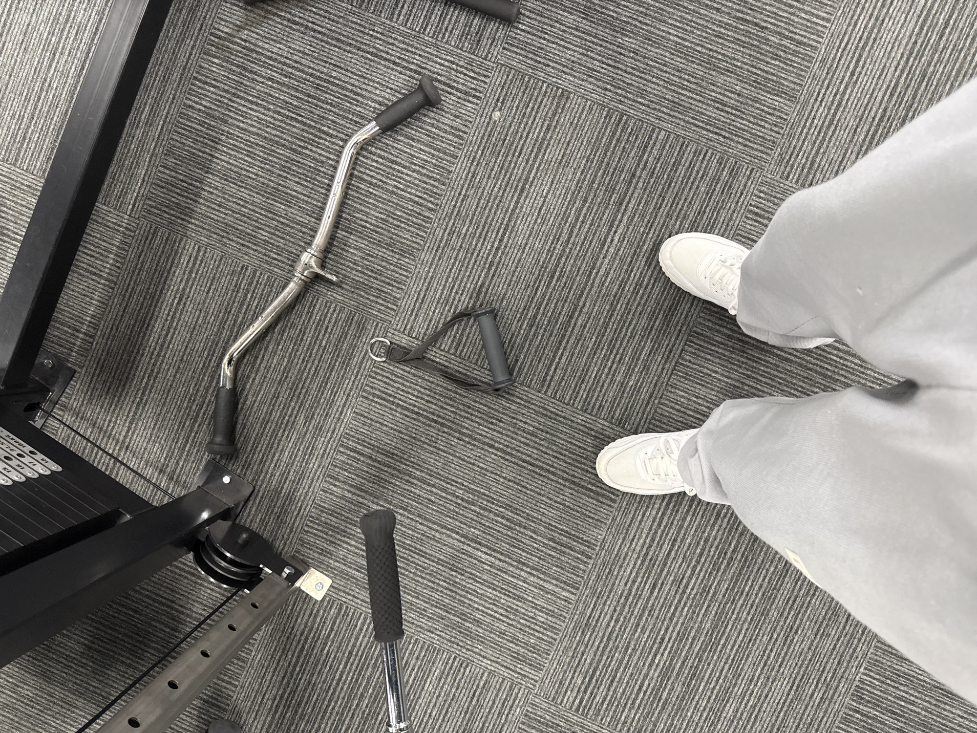 An overhead shot captures the lower body of a person standing on a patterned dark grey carpet in what appears to be a gym or fitness facility. The person is wearing light grey sweatpants and white athletic sneakers. The carpet is composed of modular tiles, featuring alternating stripe directions that create a dynamic visual texture.
To the left, a portion of a black gym cable machine is visible, including a weight stack with several plates. Numbers indicating weight levels are clearly discernible, such as "18," "16," "14," "12," "10," "8," "6," "4," "2" on one side, and "17," "15," "13," "11," "9," "7," "5," "3," "1" on the other. Pulleys and metal support frames of the machine are also part of the composition.
Two cable machine attachments lie on the carpet: a chrome-plated, curved lat pulldown or EZ curl bar with black rubber grips, and a black D-handle with a fabric strap. Another black grip on a silver bar is visible at the very bottom of the frame. The scene suggests an indoor setting, likely a gym in Letchworth, United Kingdom, where the person is either about to engage in or has just completed a workout.