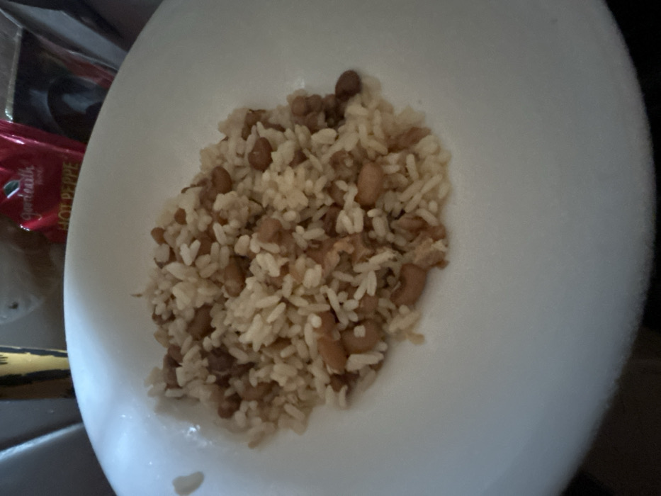 The scene, located in the city of Egbe, Nigeria, displays a white, circular bowl centrally filled with a cooked meal. The meal consists of light-colored rice mixed with numerous small, light brown beans. The bowl itself is plain white with a subtly curved rim. The background elements are out of focus. In the upper left background, a yellow and brown patterned object is partially visible. In the upper right background, a red flexible package or pouch with the visible text "Good Earth" and "HOT PEPPE" is present. No people are depicted. The lighting indicates an indoor setting.