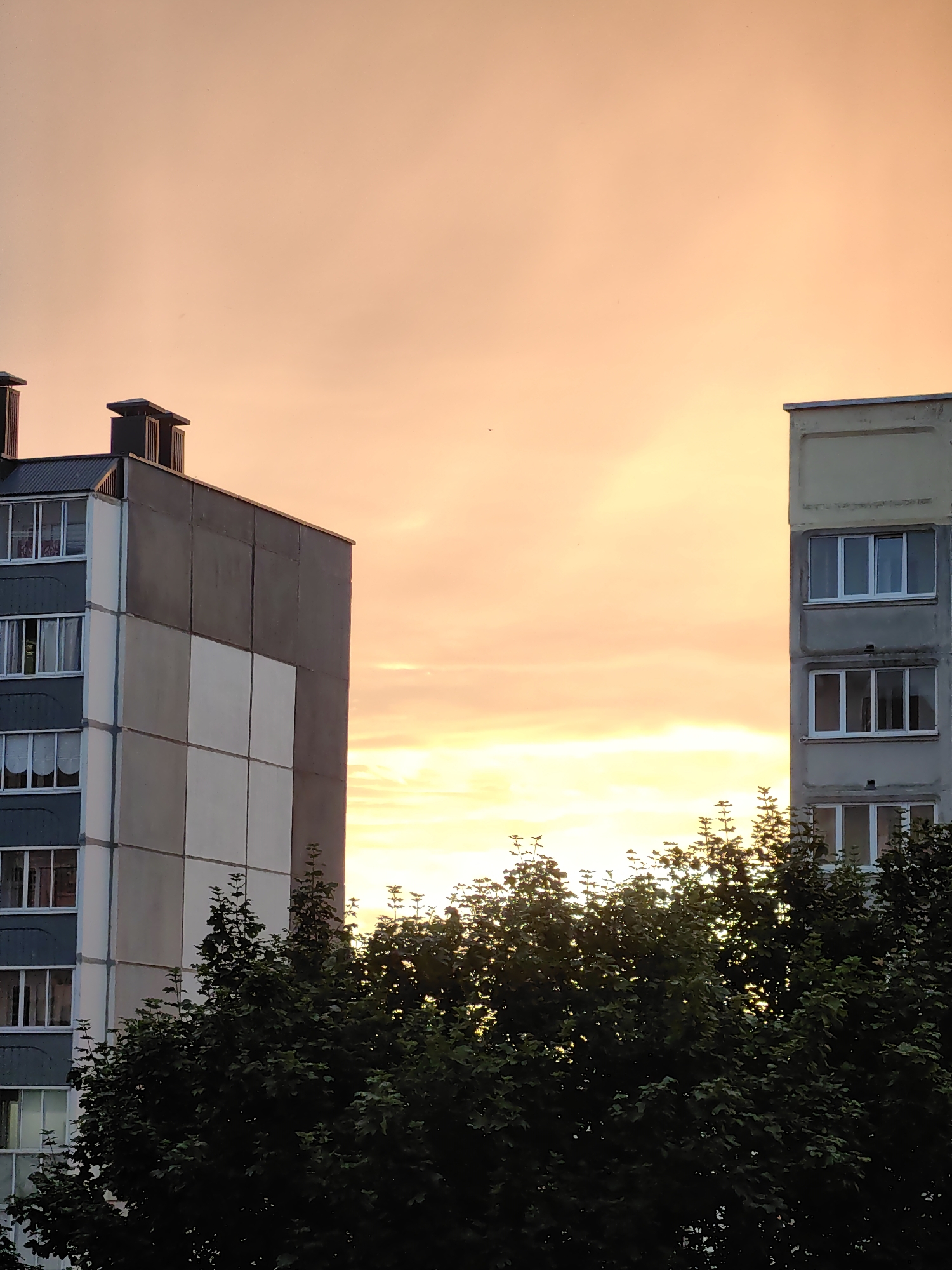 The image depicts two residential buildings against a backdrop of a sunset. The building on the left is taller and features a grey facade with contrasting white squares. It has multiple windows and two chimneys on its roof. The building on the right is lighter in color, also with several windows. Lush green trees partially obscure the lower portions of both buildings. The sky is filled with warm hues of orange and yellow, indicating the sun is setting. The overall scene suggests an urban or suburban setting in Fanipal, Belarus, during the evening. There is no visible text in the image.