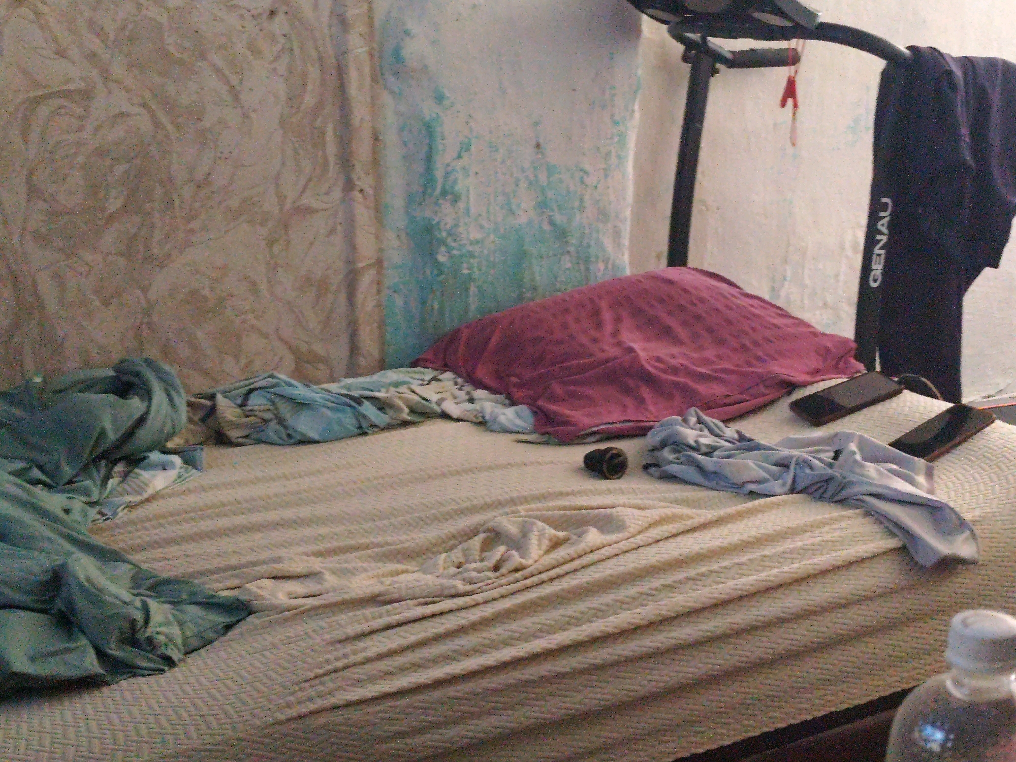 The image depicts a messy bedroom, possibly in an apartment, in the city of Balqaş, Kazakhstan. The room is characterized by a disheveled bed with several pieces of clothing strewn across it, including a pink blanket or throw, blue and white patterned sheets, and a pair of dark blue sweatpants with "GENAU" written on them hanging from a black exercise machine. Two smartphones are visible on the bed, one with a red case. The walls appear to be textured and are painted with a patchy white and blue design. In the foreground, a clear plastic bottle with a white cap is visible, suggesting it contains liquid. The overall impression is one of untidiness and everyday life.