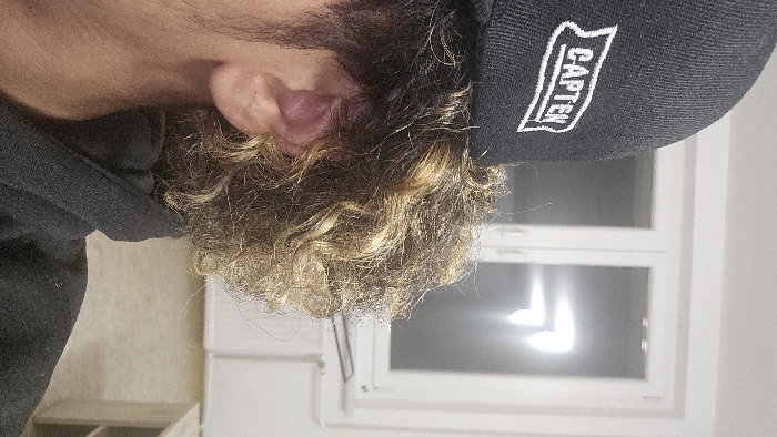 A person's head and part of their shoulder are visible. The individual has curly, highlighted hair, with dark roots and blonde tips. They are wearing a black hooded sweatshirt and a black baseball cap with white lettering, "CAPTEN" on the side. The background includes a white wall and a window with bright light shining through. A white cabinet or shelf is partially visible in the lower left corner. The image appears to be taken from a low angle.