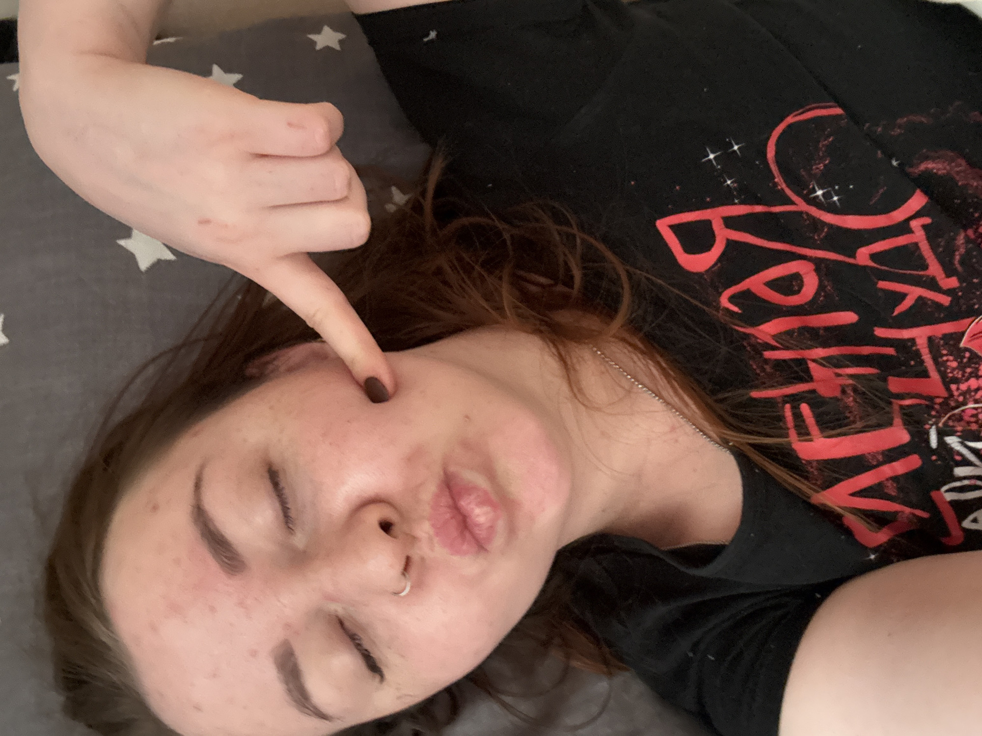 A young woman lies on a gray bedsheet patterned with white stars. She is wearing a black t-shirt with red lettering. Her eyes are closed, and her mouth is puckered. She has a nose ring in her left nostril. Her right hand is near her face, with a finger pointing at her right cheek. Her hair is dark brown. There is a dark spot on her right cheek. The location in the city of Izhevsk, Russia cannot be determined from this image.
