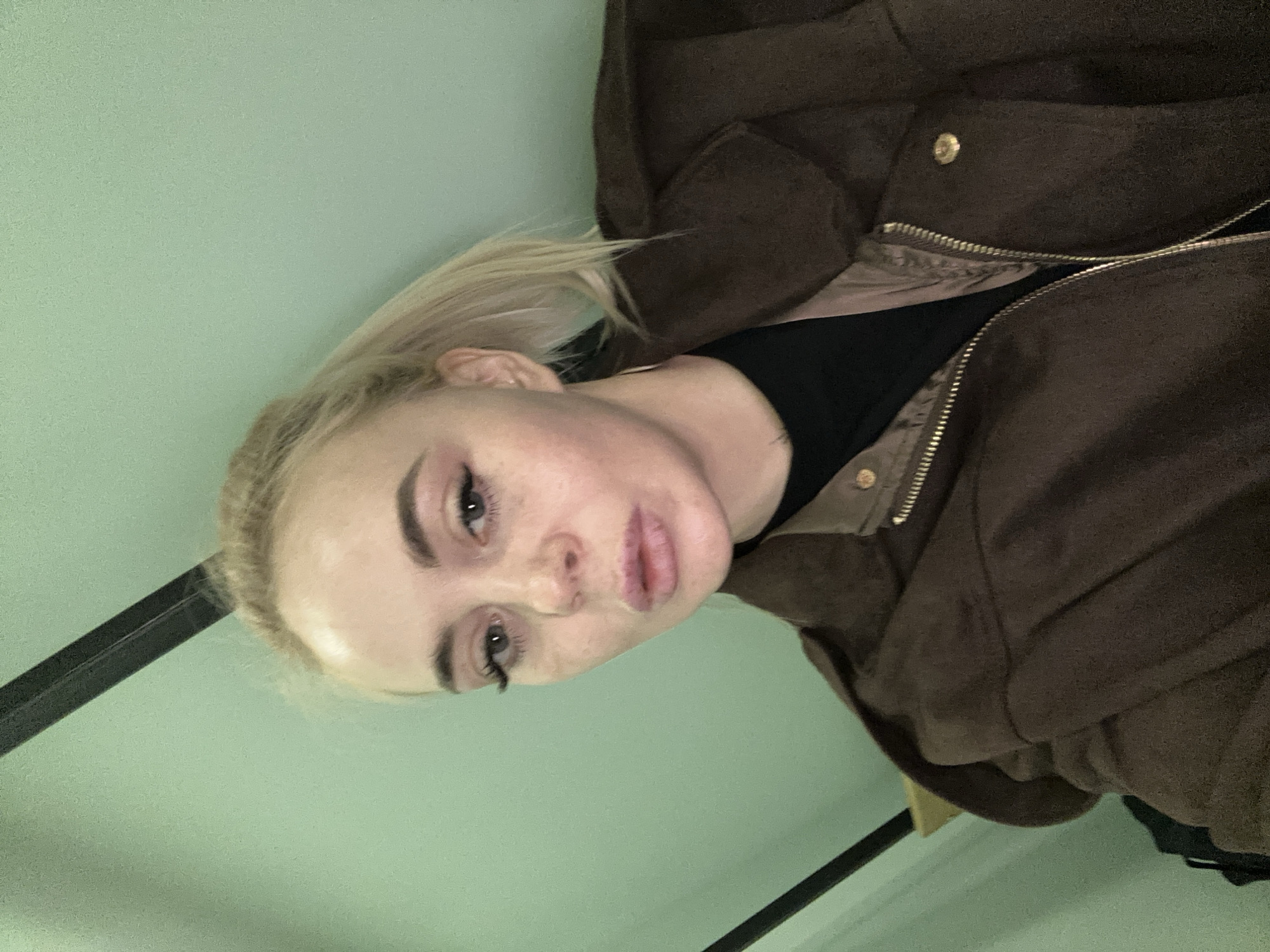 A fair-skinned, blonde-haired woman is visible in the image. She is wearing a black shirt and a brown jacket. The background is a light green wall. A black metal bar is visible in the lower-left corner. The location is not evident from the image.