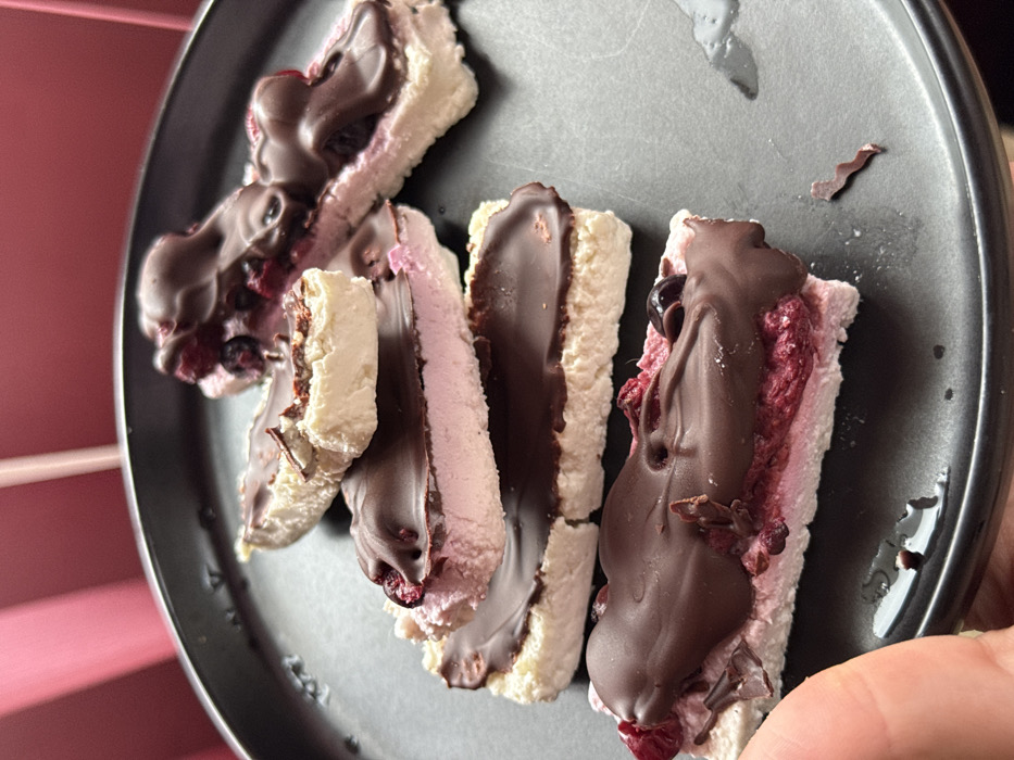 The image displays a close-up of multiple layered dessert bars arranged on a dark, matte, circular plate. A human thumb and forefinger are visible in the lower-left quadrant, supporting the plate. Each bar features an off-white, textured base layer, topped by a distinct pinkish-red layer containing fragmented dark red and dark blue berries. The uppermost layer consists of a thick, melted dark chocolate coating, with small, irregular pieces, resembling dried fruit or chocolate flakes, embedded within and on top of it. Scattered droplets of liquid and a single small, wavy, dark brown fragment are present on the plate surface. The background is a blurred field of dark red, exhibiting subtle vertical striations. The scene is located in the city of Galabovtsi, Bulgaria.